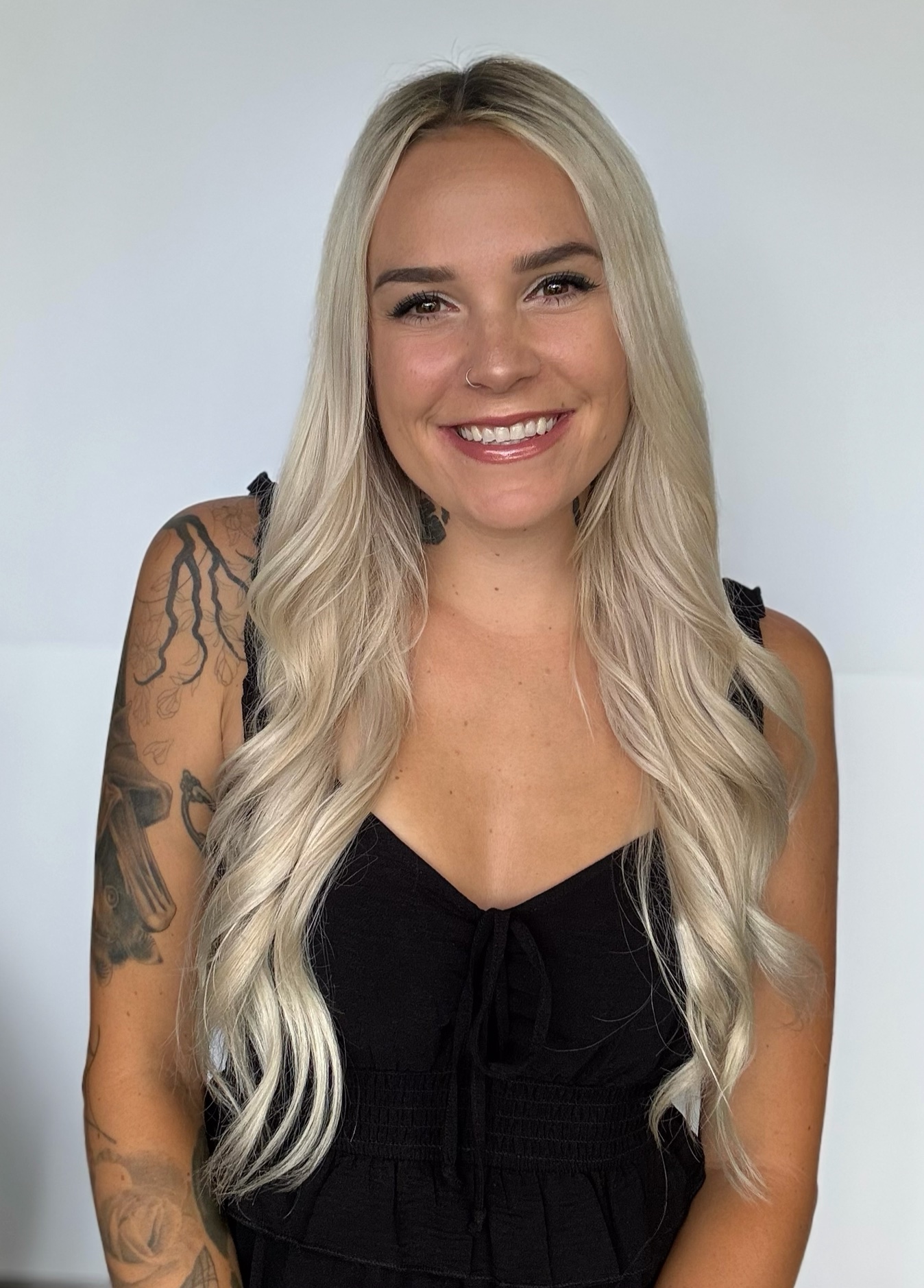 Young woman with long, wavy blonde hair smiling, wearing a black sleeveless top, tattoos on her left arm, against a plain background.