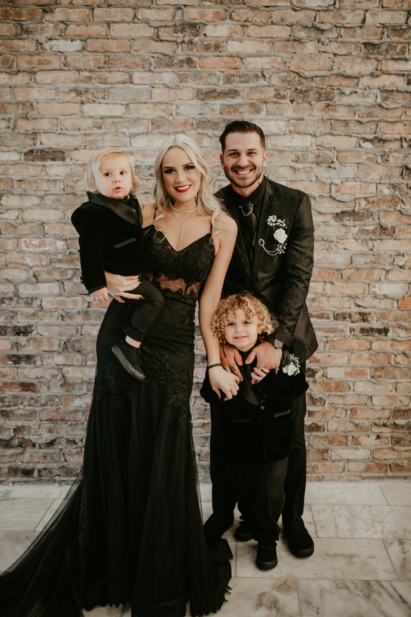 Group of four people standing in front of a brick wall, smiling, with a woman in a black dress, a man, and two children.