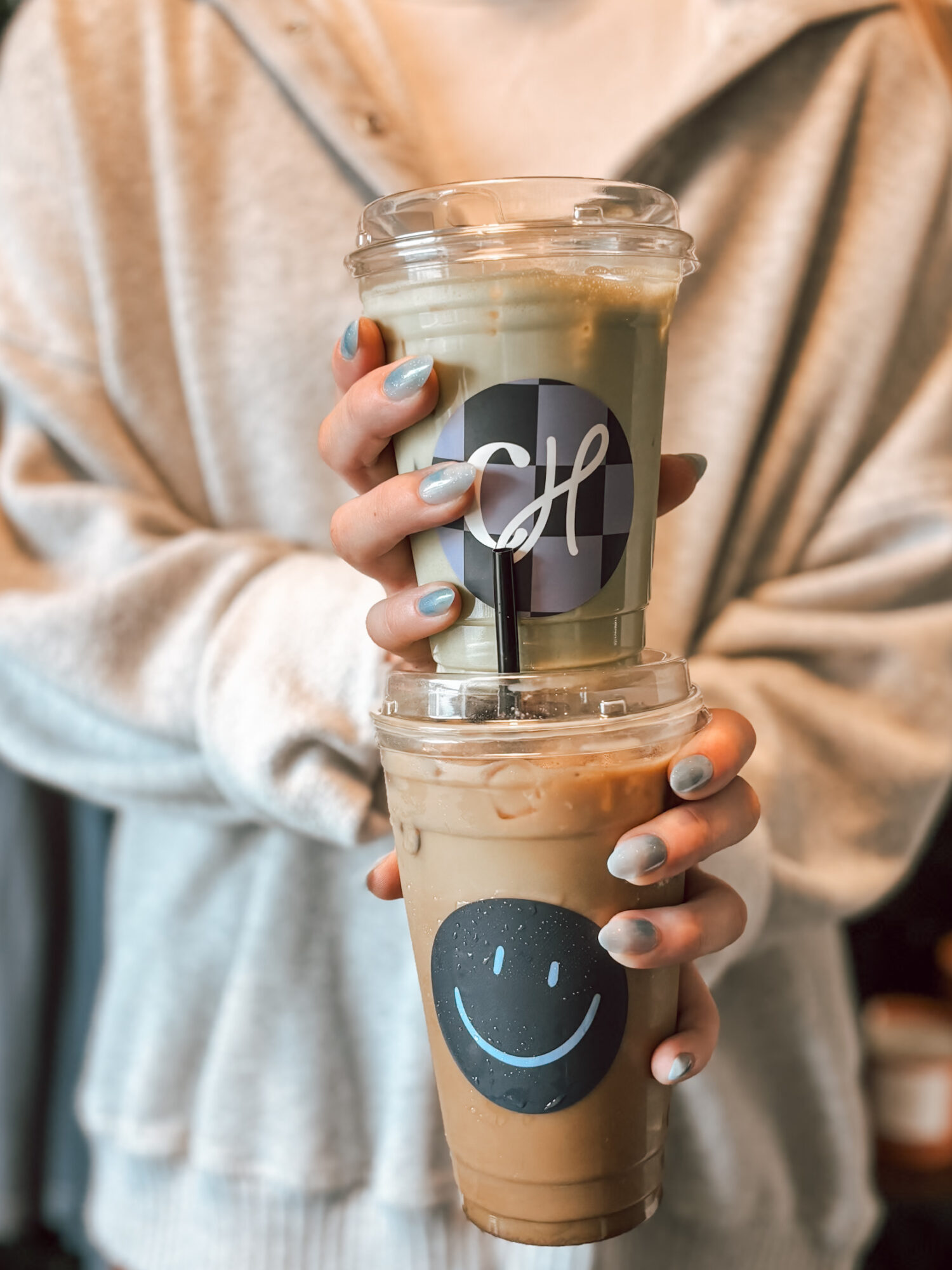 Person holding two iced coffee drinks with smiley face and logo, wearing a beige hoodie, nails painted blue.