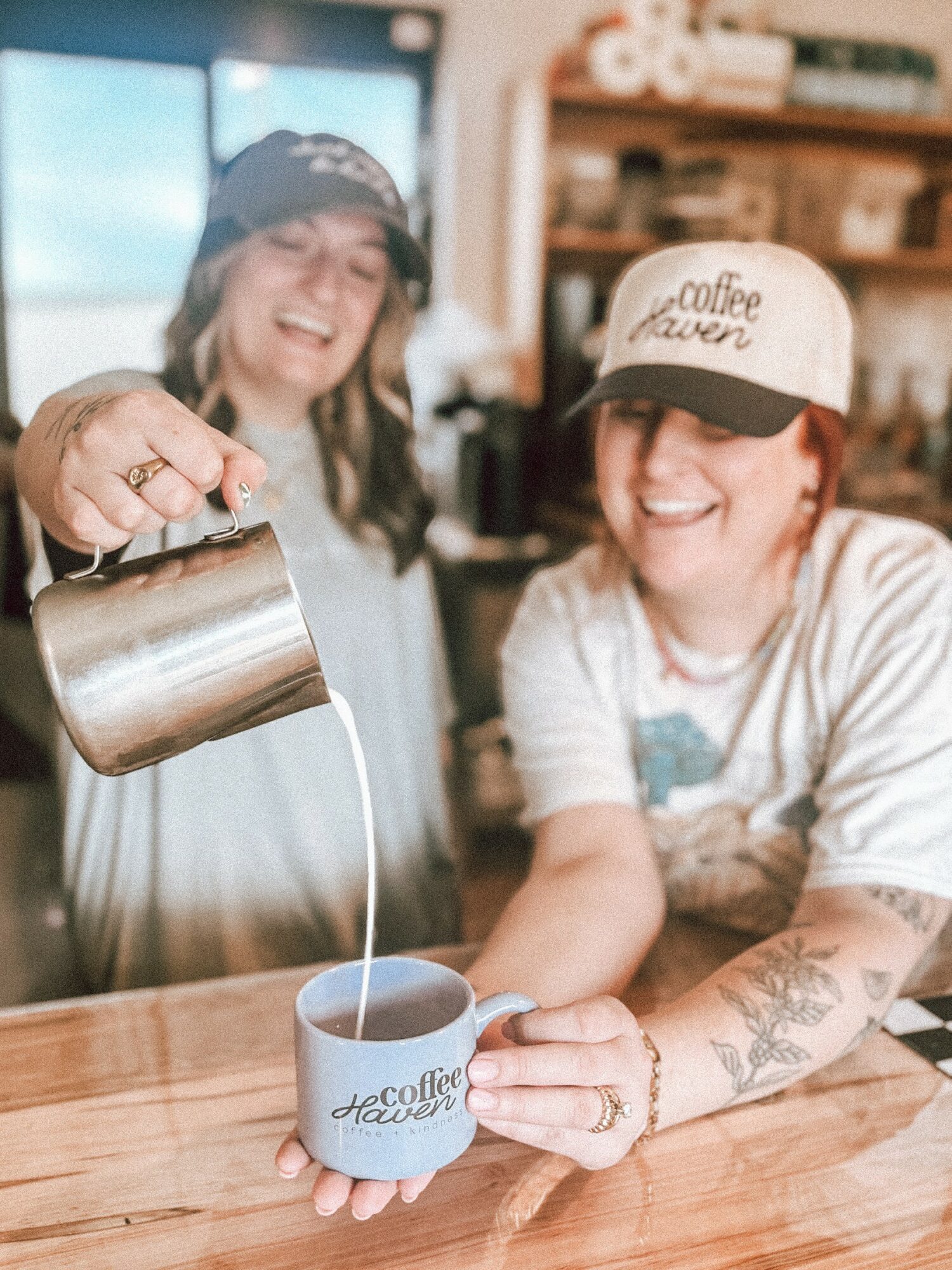 Two women smiling, one pouring milk into a coffee mug, in a cozy cafe setting.