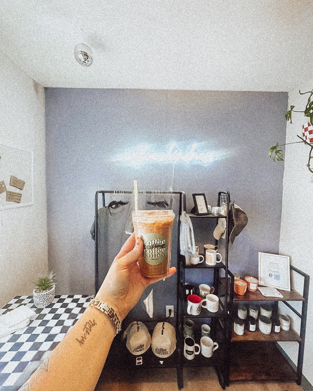 Person holding a coffee cup in a room with shelves and a checkered table, wall with a neon sign, and plants.