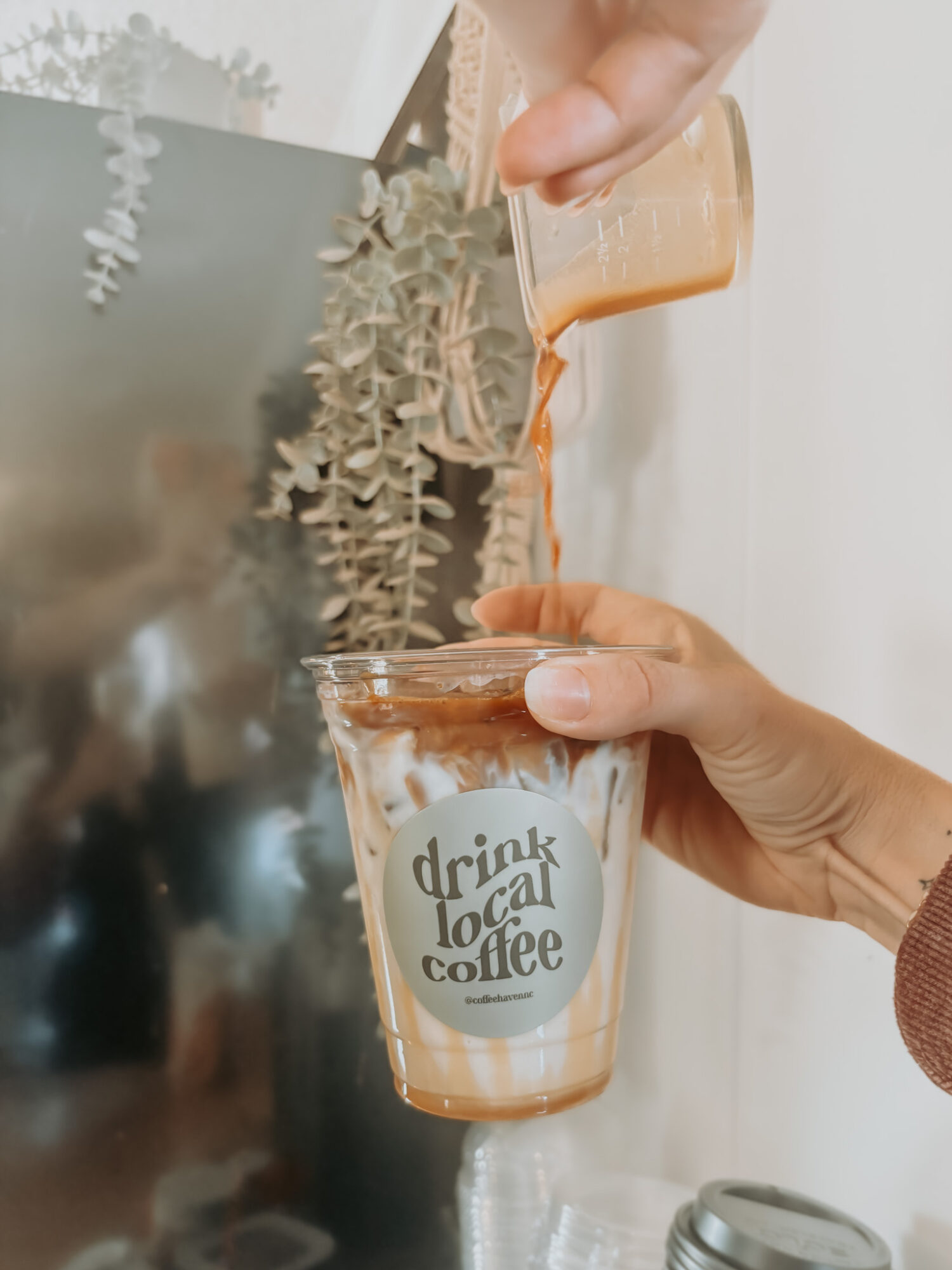 Person pouring coffee into a glass with a logo, with plants in the background.