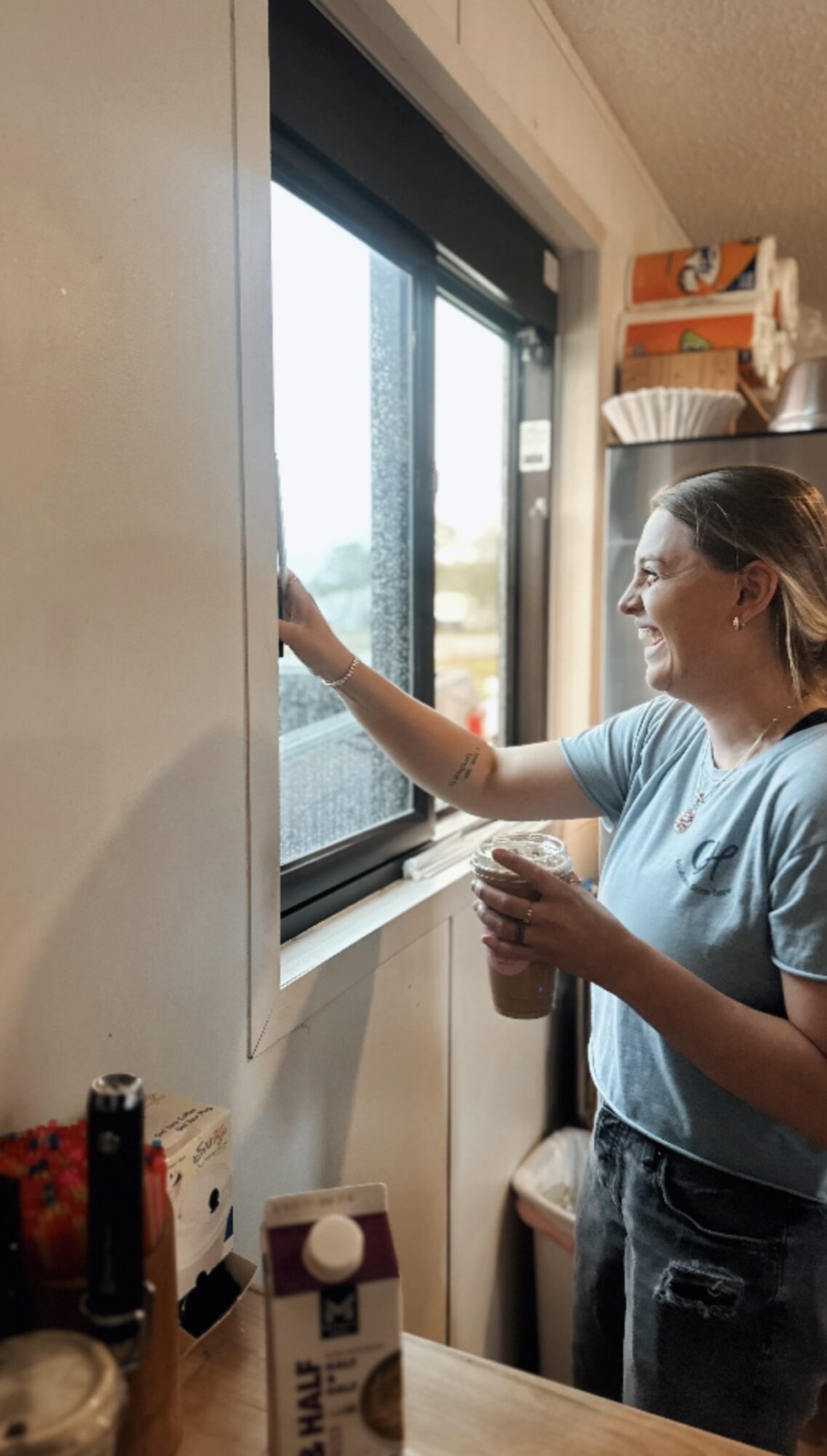 Woman cleaning window with spray bottle inside a room, sunlight outside, shelves and items on wall, countertop with objects.