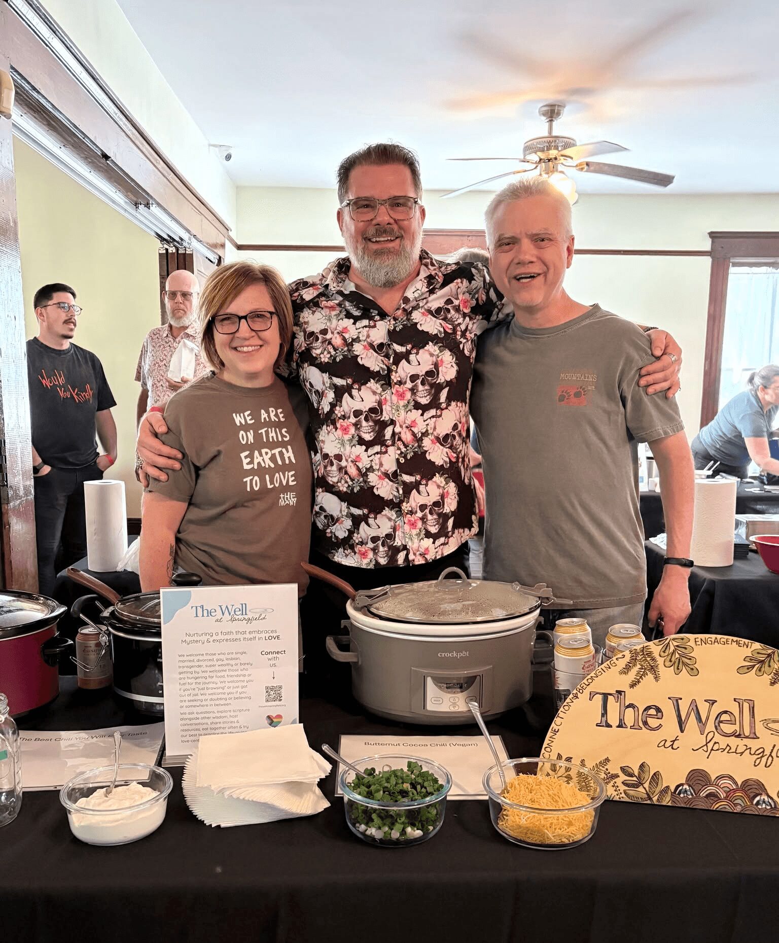 Three people stand behind a table with food and signs, smiling indoors with other people in the background.