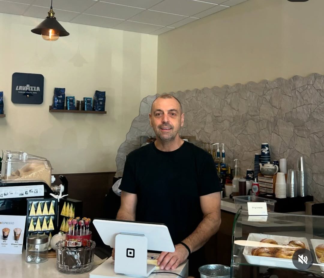 Man standing behind a counter in a cafe, smiling, with coffee cups and supplies around him.