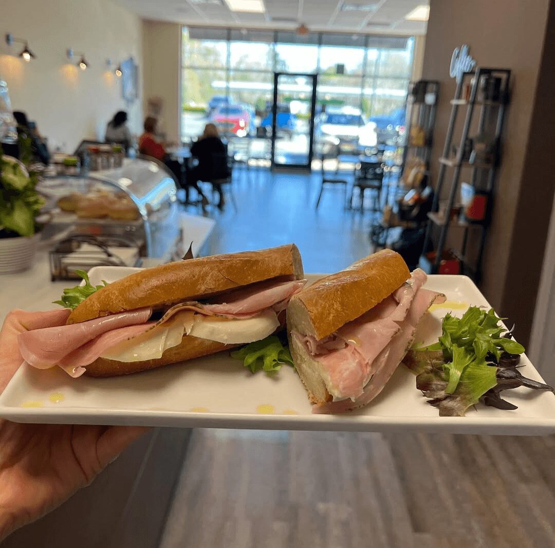 Two sandwiches with ham, cheese, and lettuce on a white plate inside a cafe, with seating and large windows in the background.