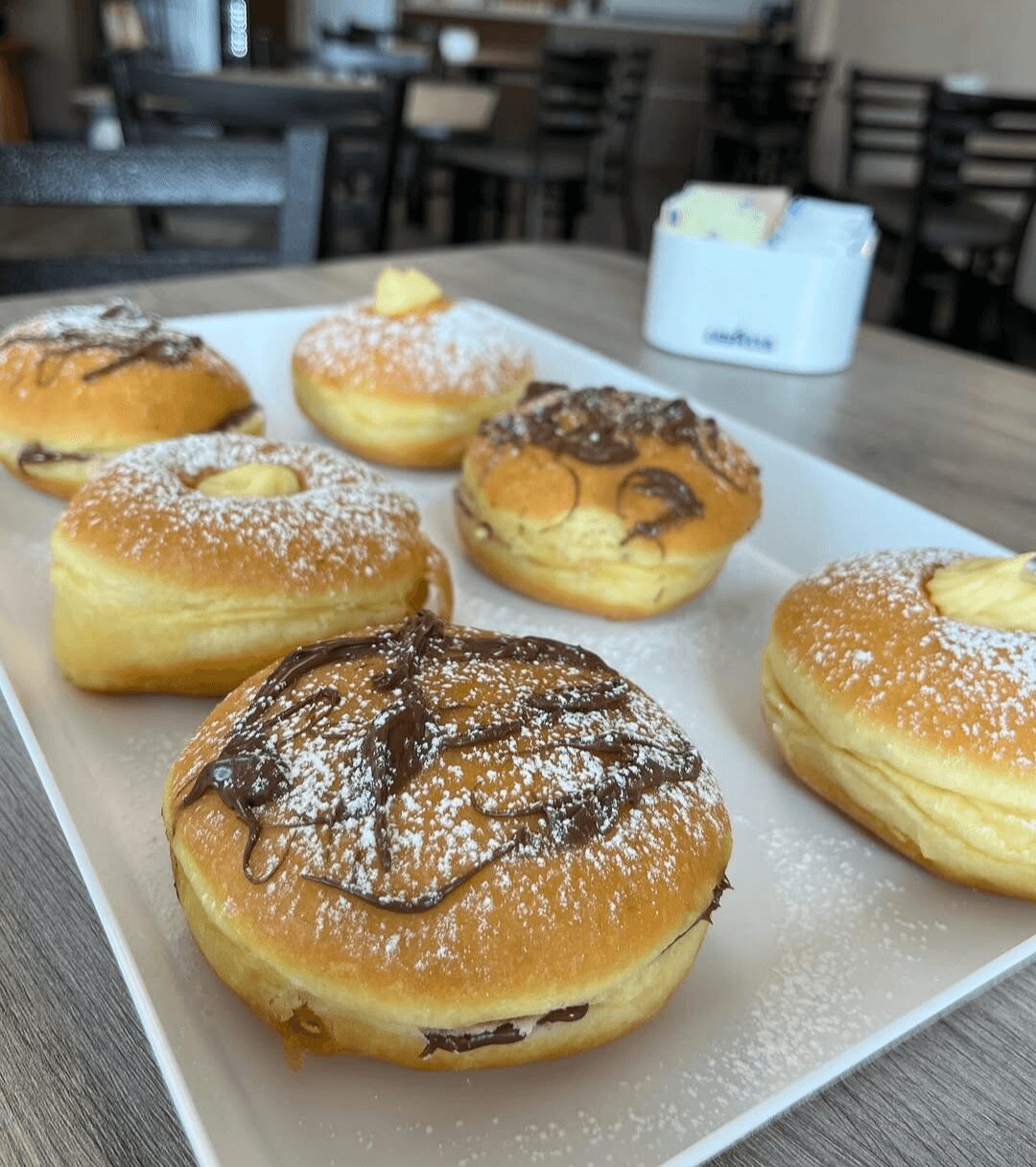 Six assorted donuts on a white tray in a cafe, with a background of tables and chairs.