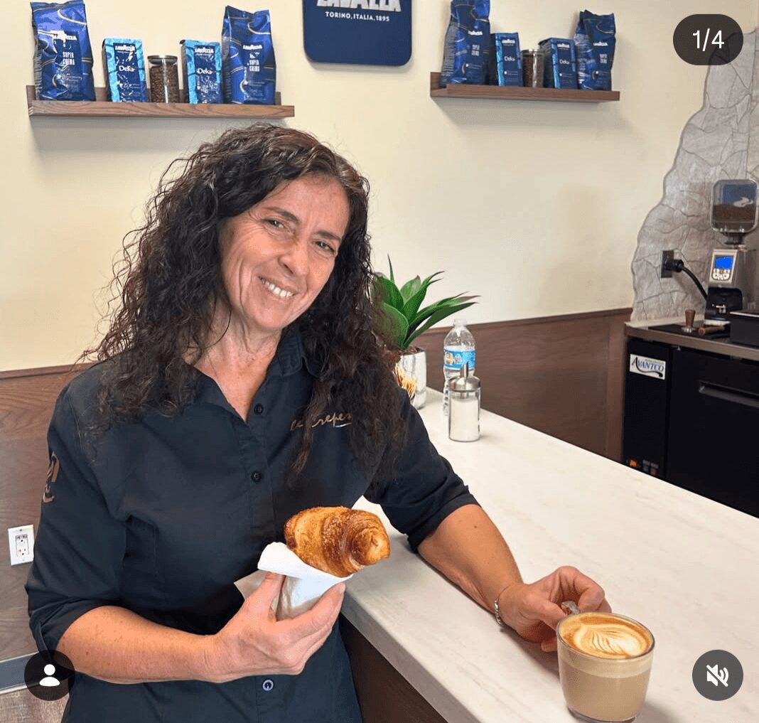 Woman with curly hair smiling, holding a croissant on a napkin, standing at a counter with a latte, in a coffee shop.