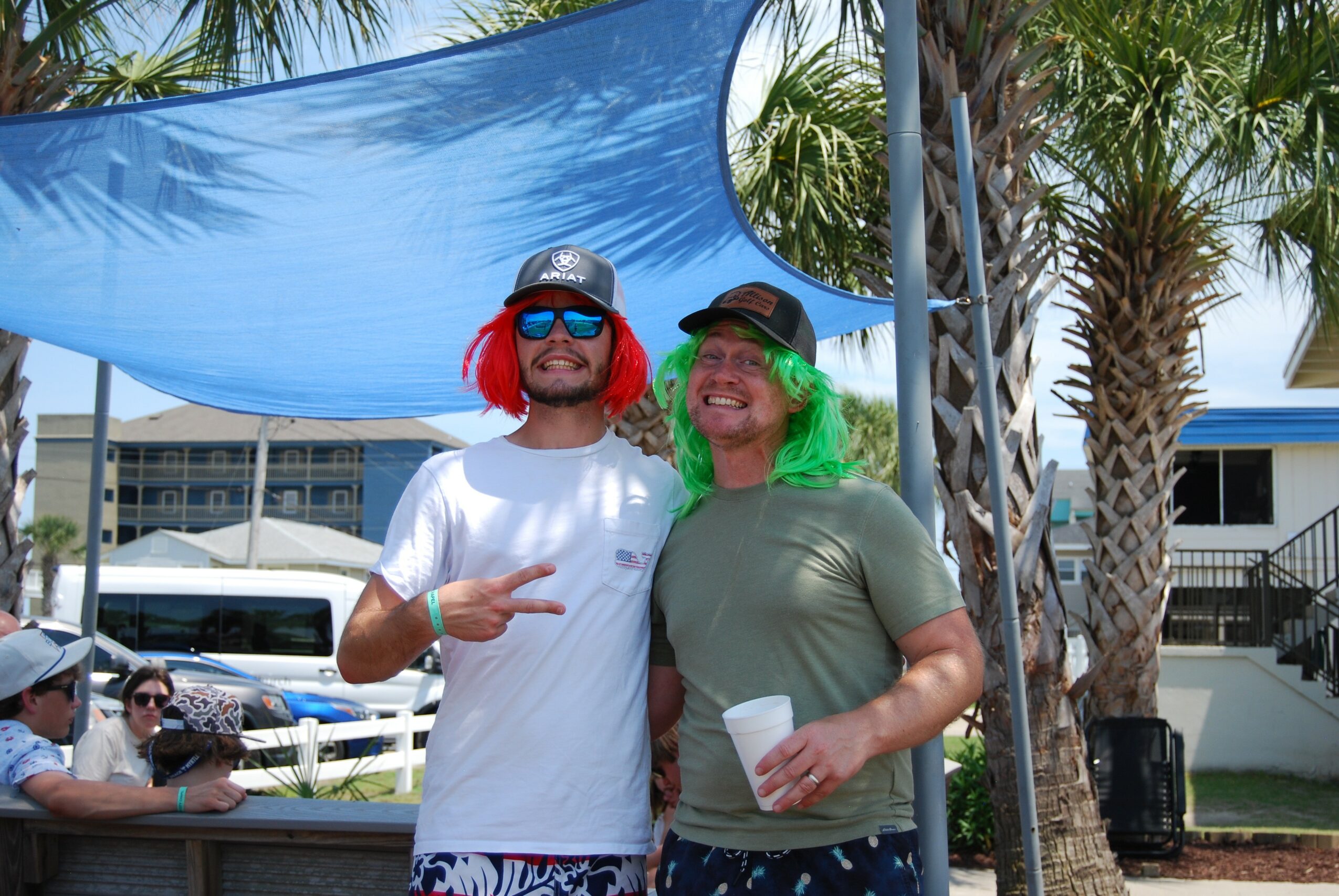 Two people with colorful wigs and sunglasses smiling outdoors, holding drinks, with palm trees and a blue canopy in the background.
