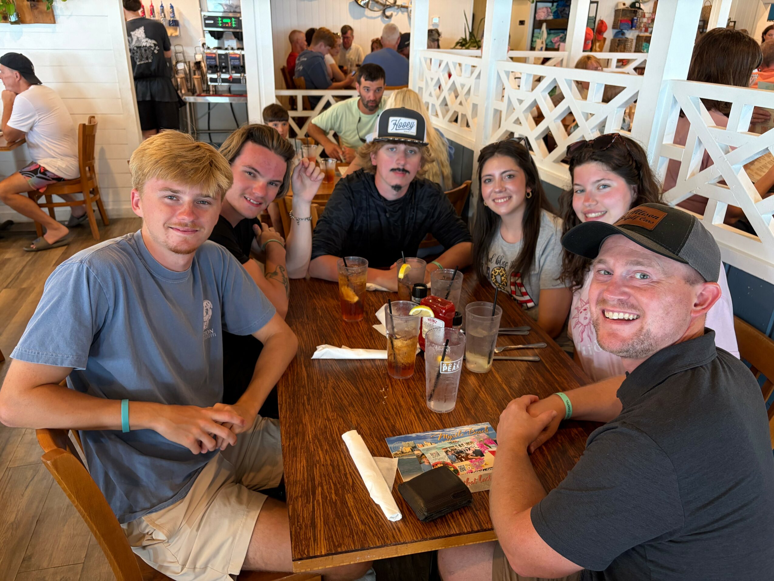 Group of six people sitting at a restaurant table, smiling, with drinks and menus, in a lively dining area.