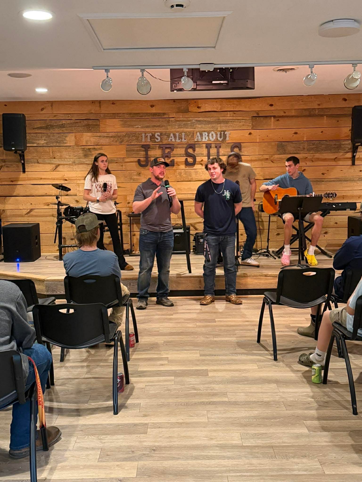 Group of five people on stage, two singing into microphones, others playing guitar and keyboard, audience seated, wooden backdrop.