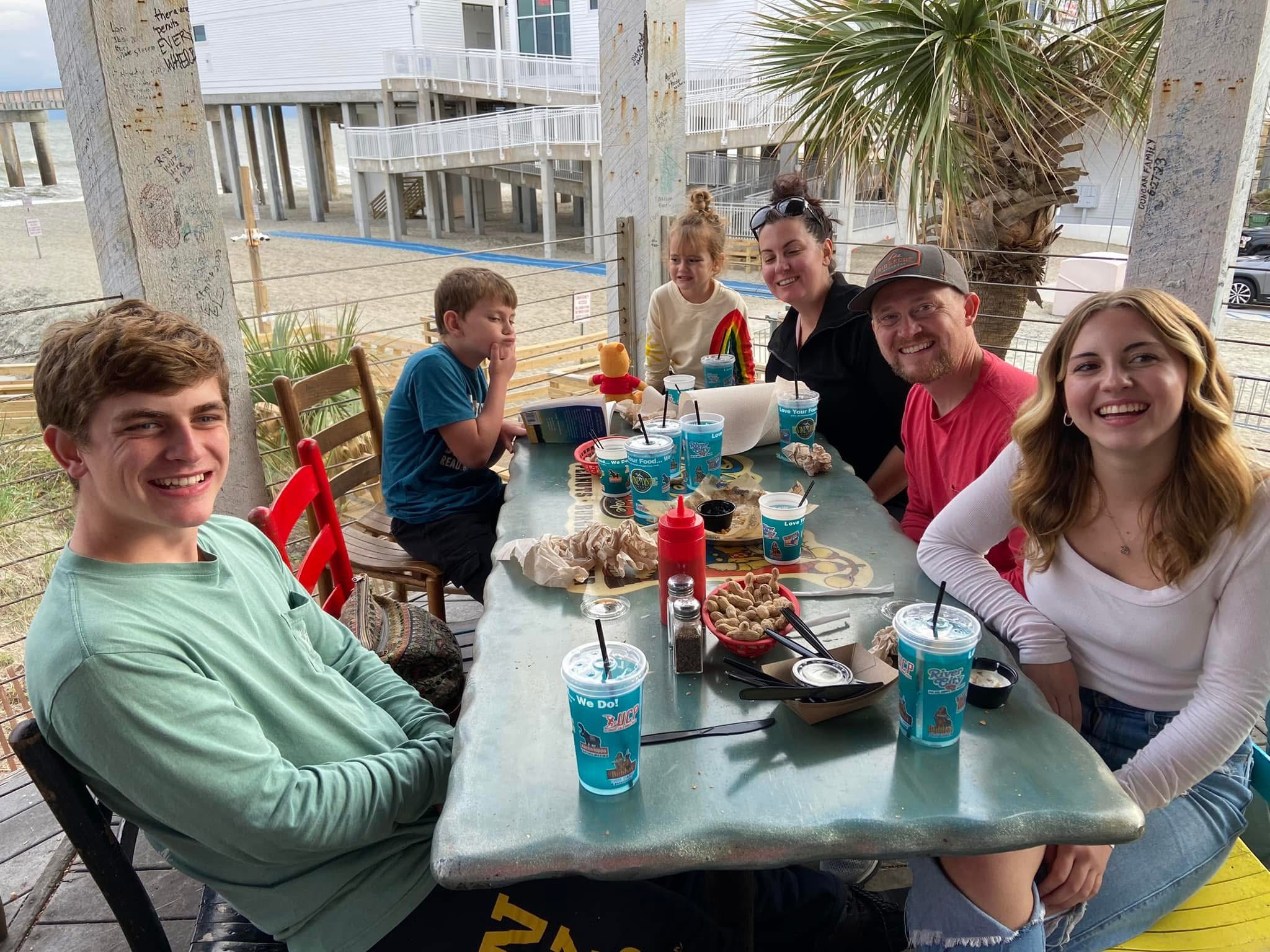 Group of six people sitting around a table outdoors, smiling, with drinks and snacks, in a sunny setting.