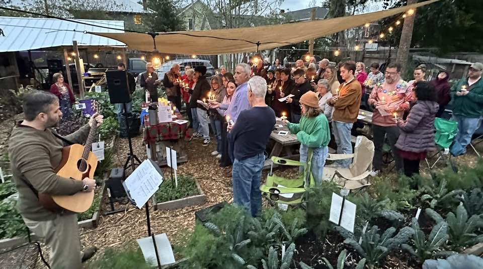 Group of people gathered outdoors under string lights, listening to a person playing guitar, with plants and tables nearby.