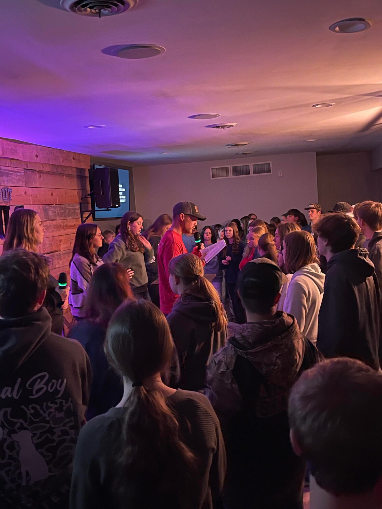 Group of people gathered in a dimly lit room with purple and orange lighting, some facing a screen on the wall.