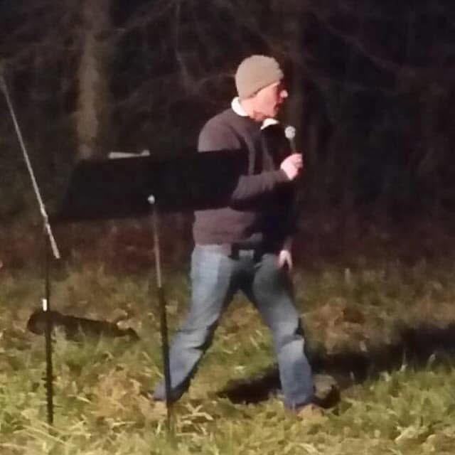 Person wearing a beanie and dark clothing holding a microphone outdoors at night, standing on grass with trees in background.
