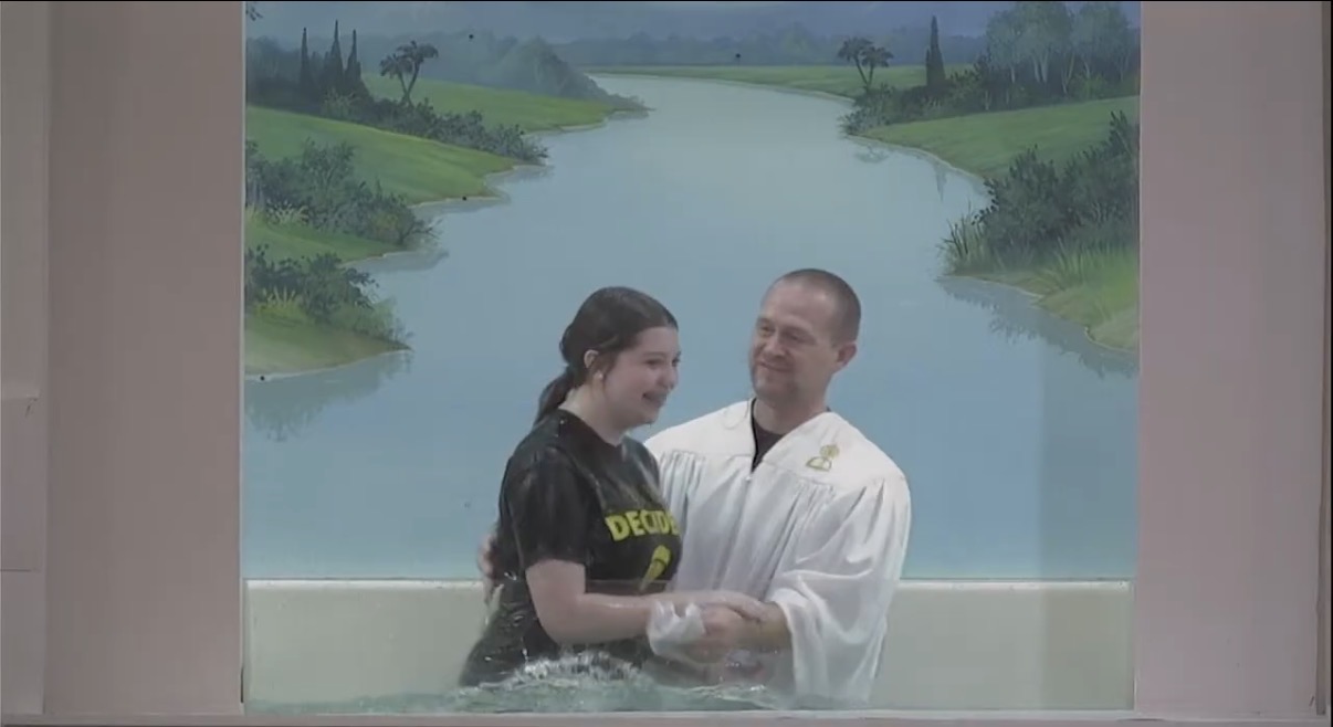 Two people standing in water with a painted landscape background, one holding a bowl, smiling at each other.