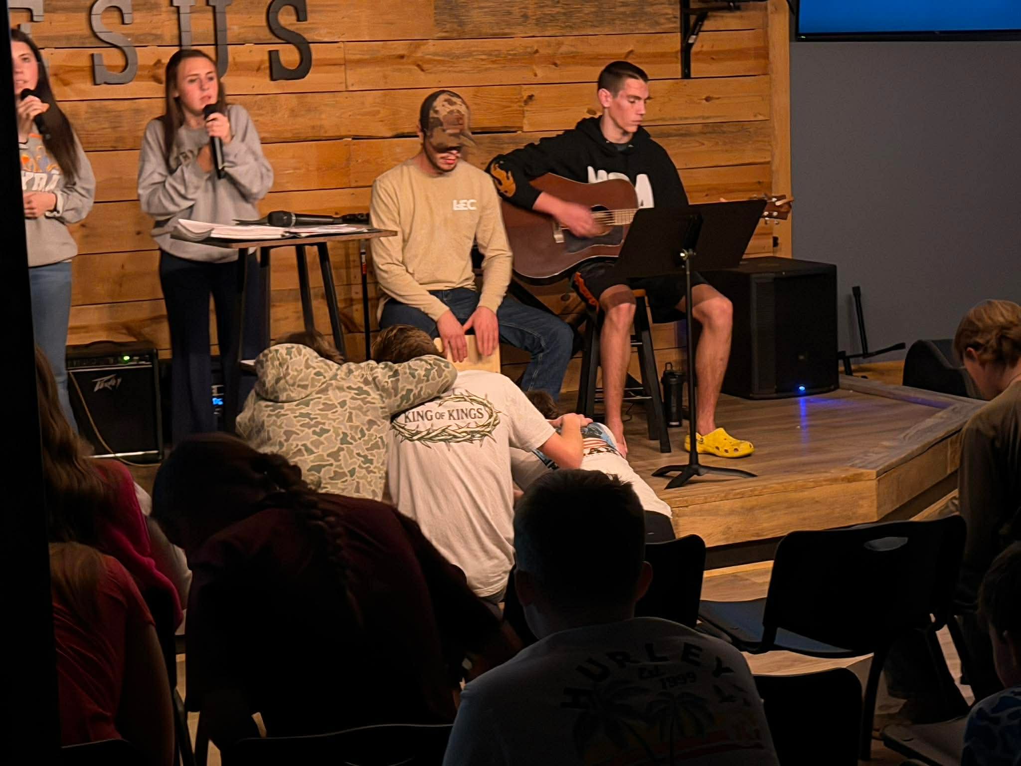 People on stage performing with a person playing guitar, audience seated watching, wooden wall background, stage setup.