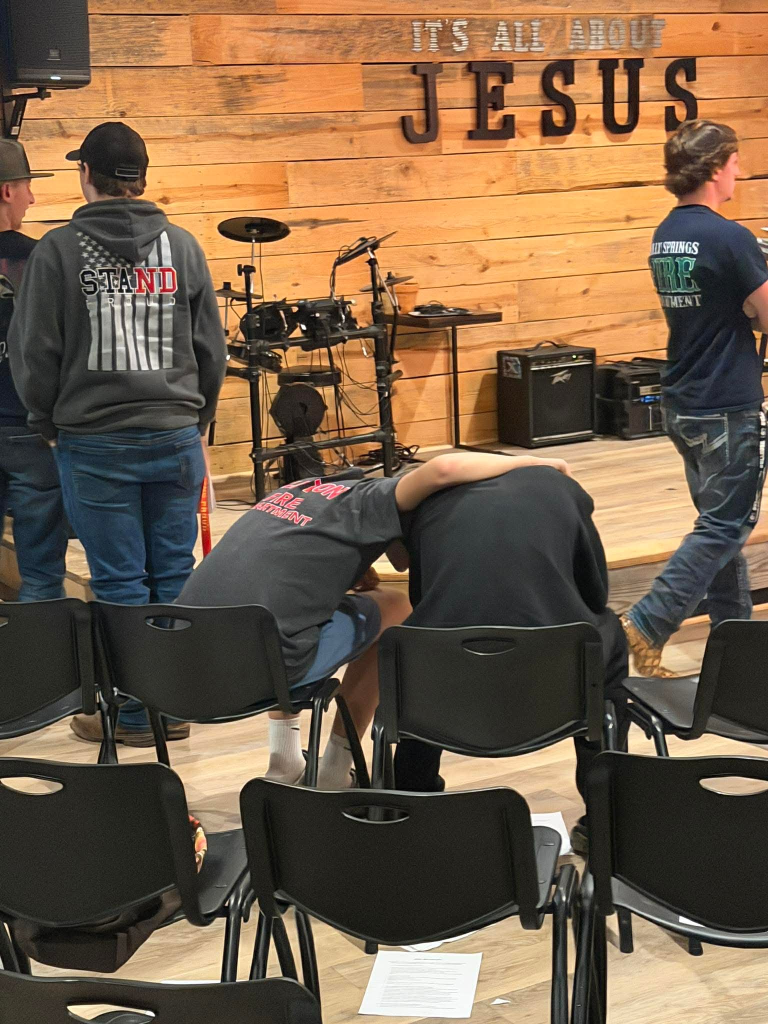 Two people sit on chairs facing a stage with musical instruments, others stand nearby, wooden wall with 'JESUS' sign in background.