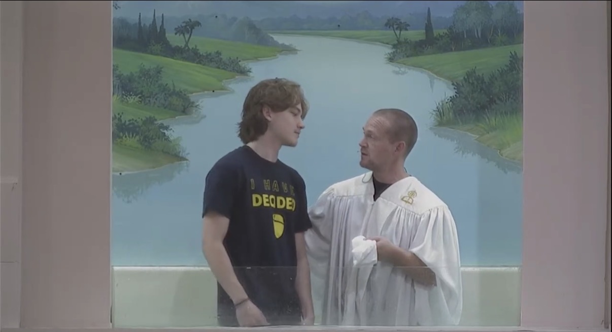 Two people standing and talking in front of a painted landscape background, one wearing a dark shirt and the other in a white gown.