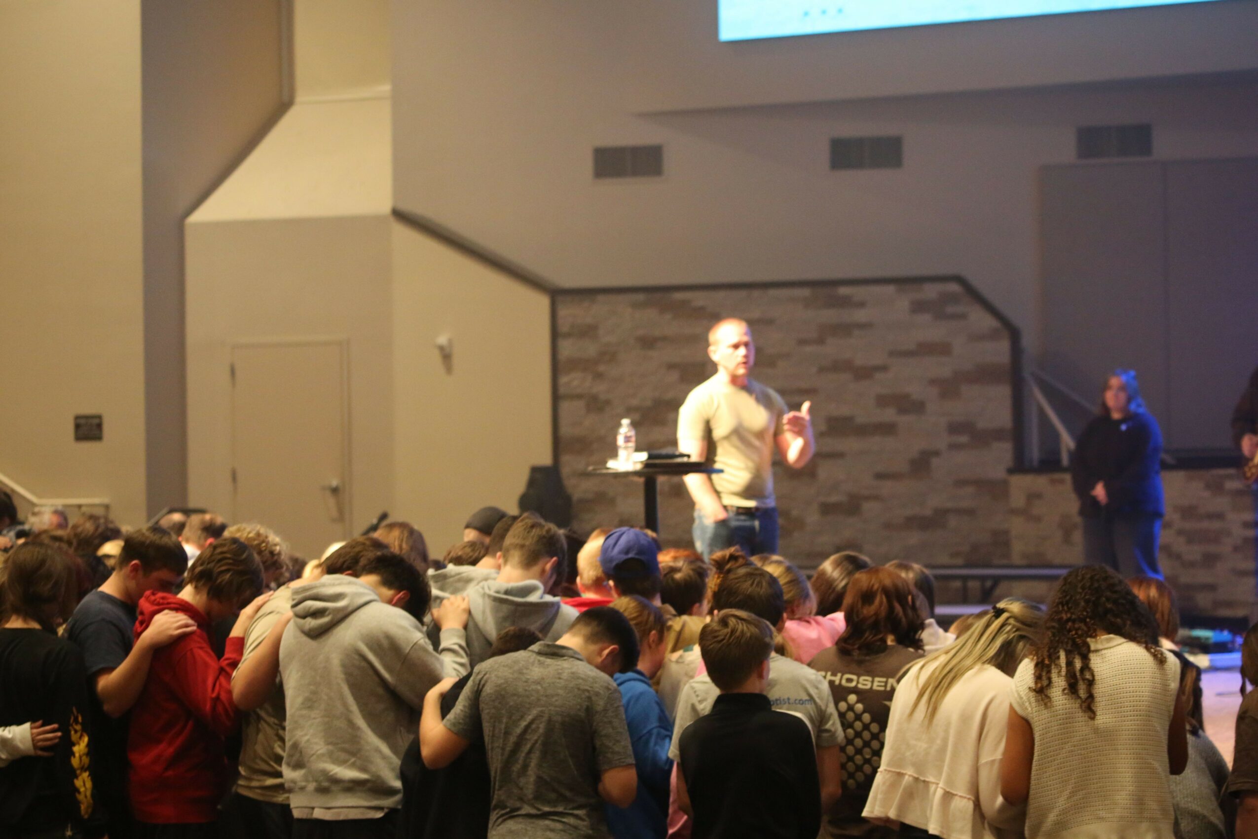 A speaker stands on stage addressing a large audience in a room with a brick wall background, some audience members are taking photos.