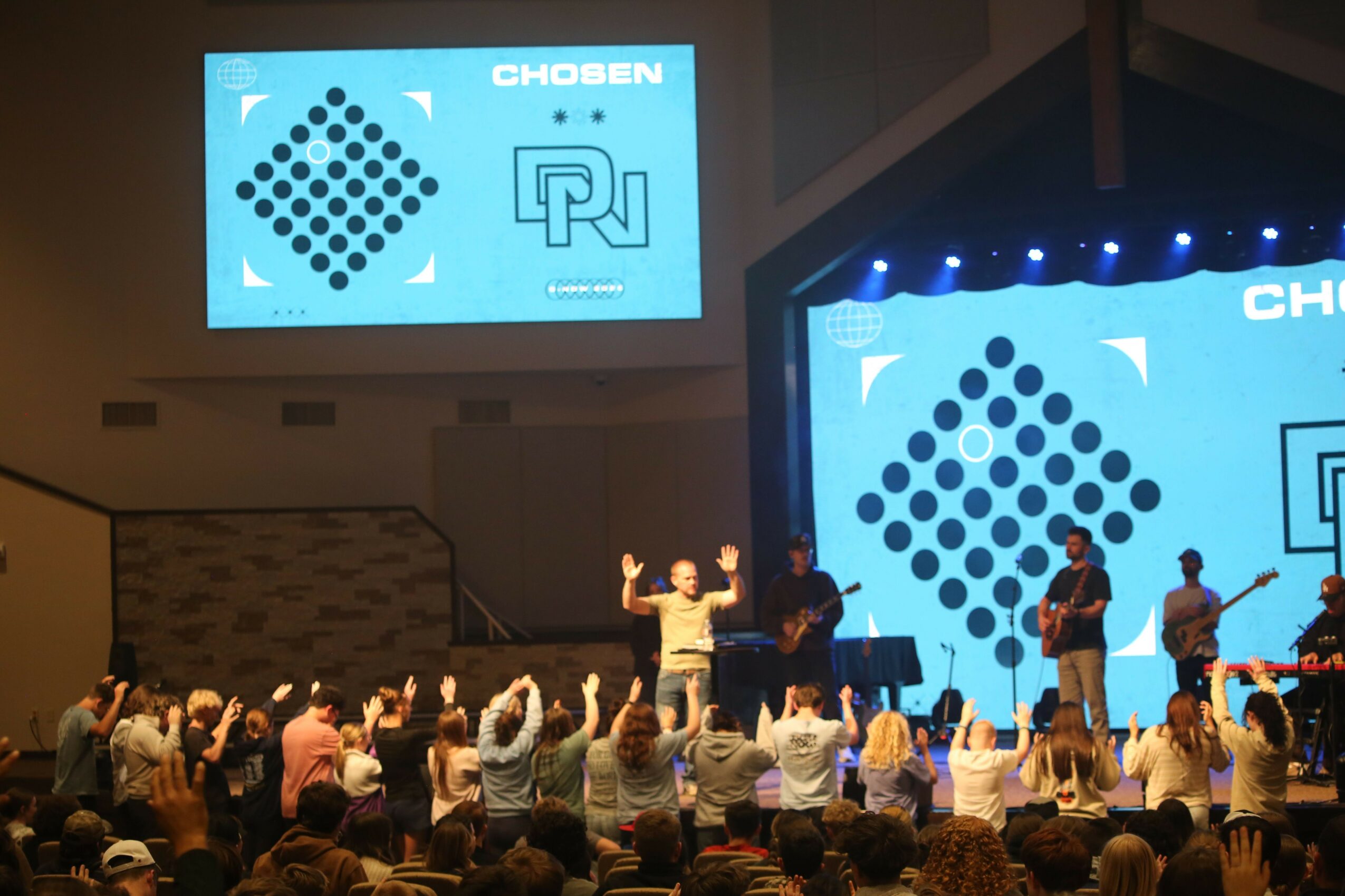 People on stage with large screens behind them displaying geometric logos, audience raising hands, indoor event setting.