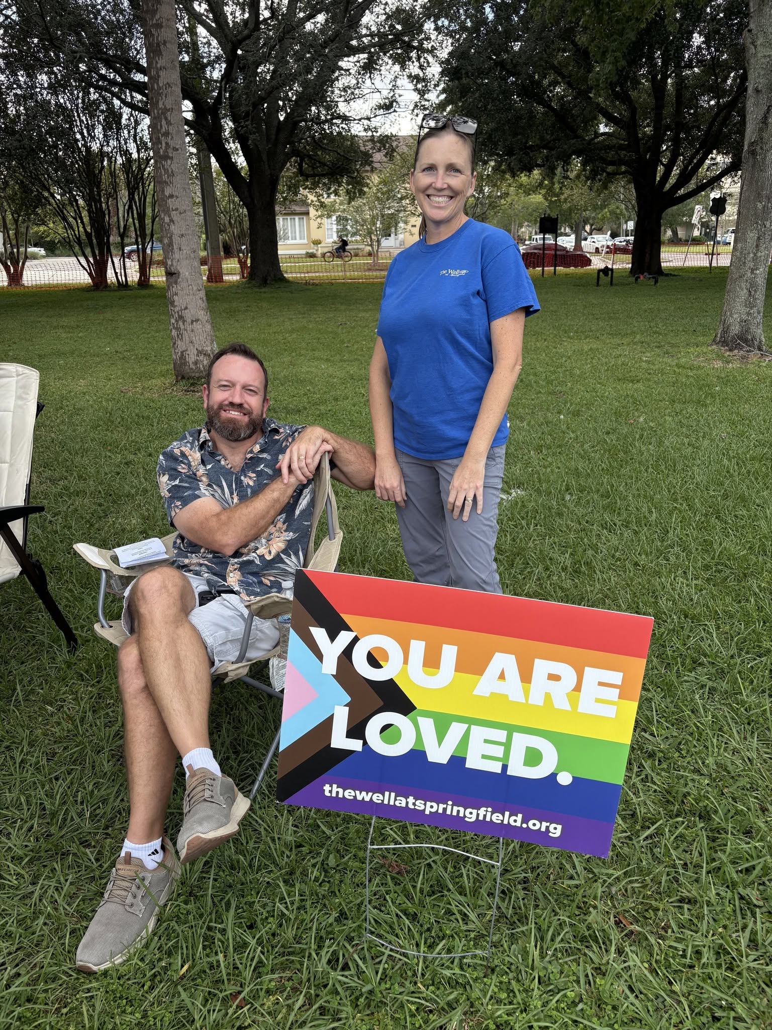 Two people outdoors with a rainbow-colored sign reading