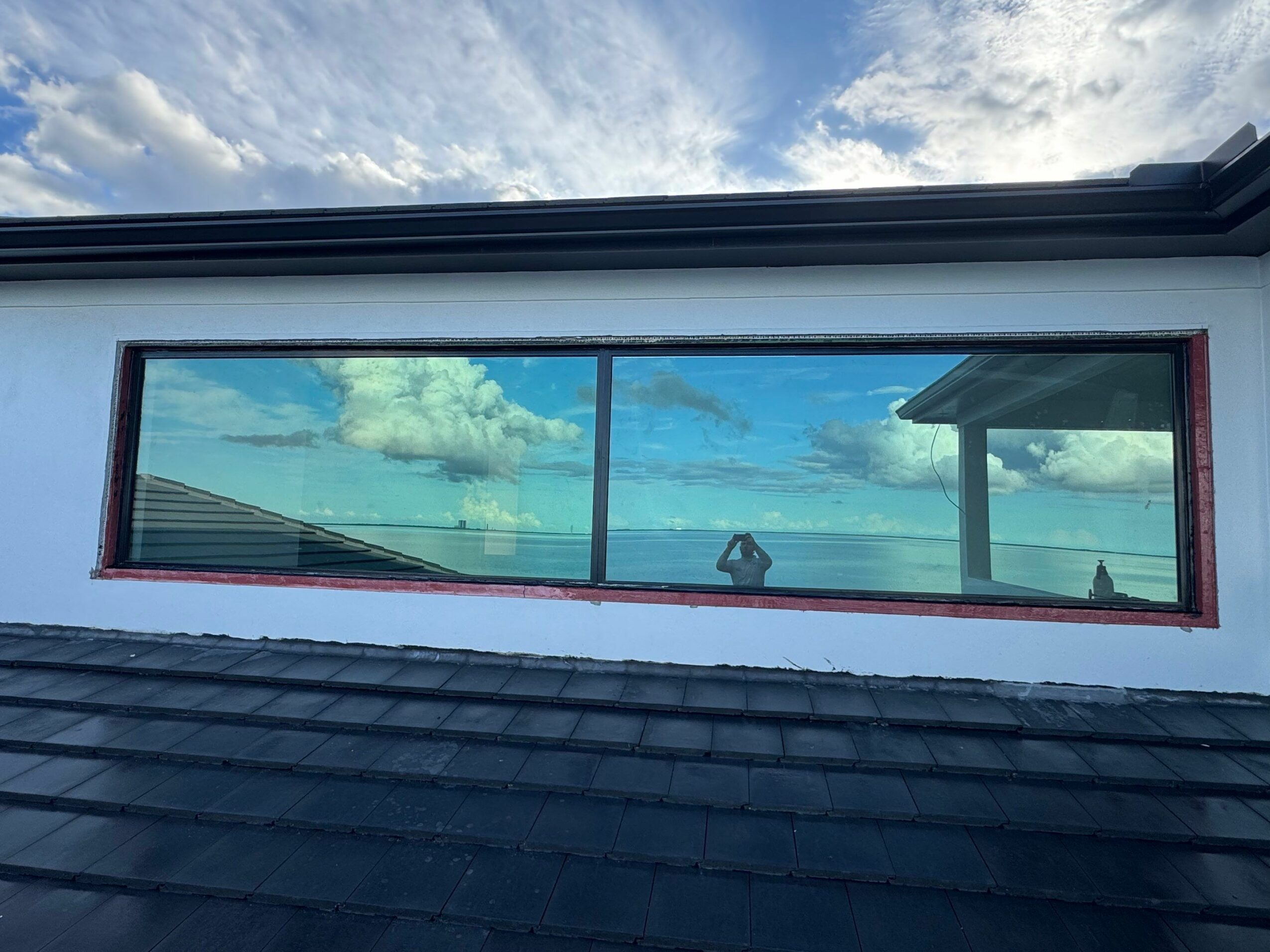 A large horizontal window reflects a cloudy sky and part of a roof, framed by a white wall with red trim.