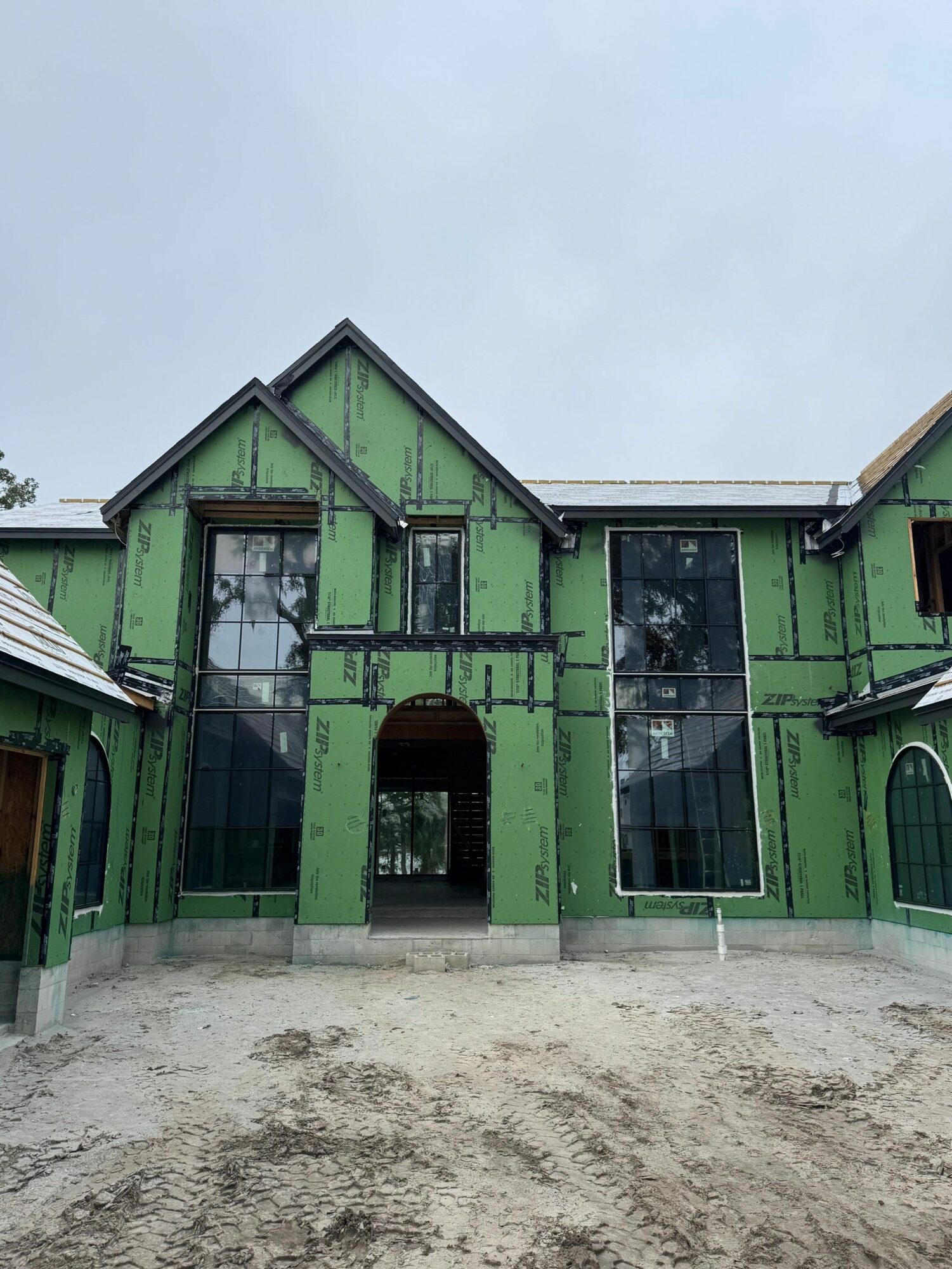 Green house under construction with large windows and an arched entrance, on a dirt ground, cloudy sky above.