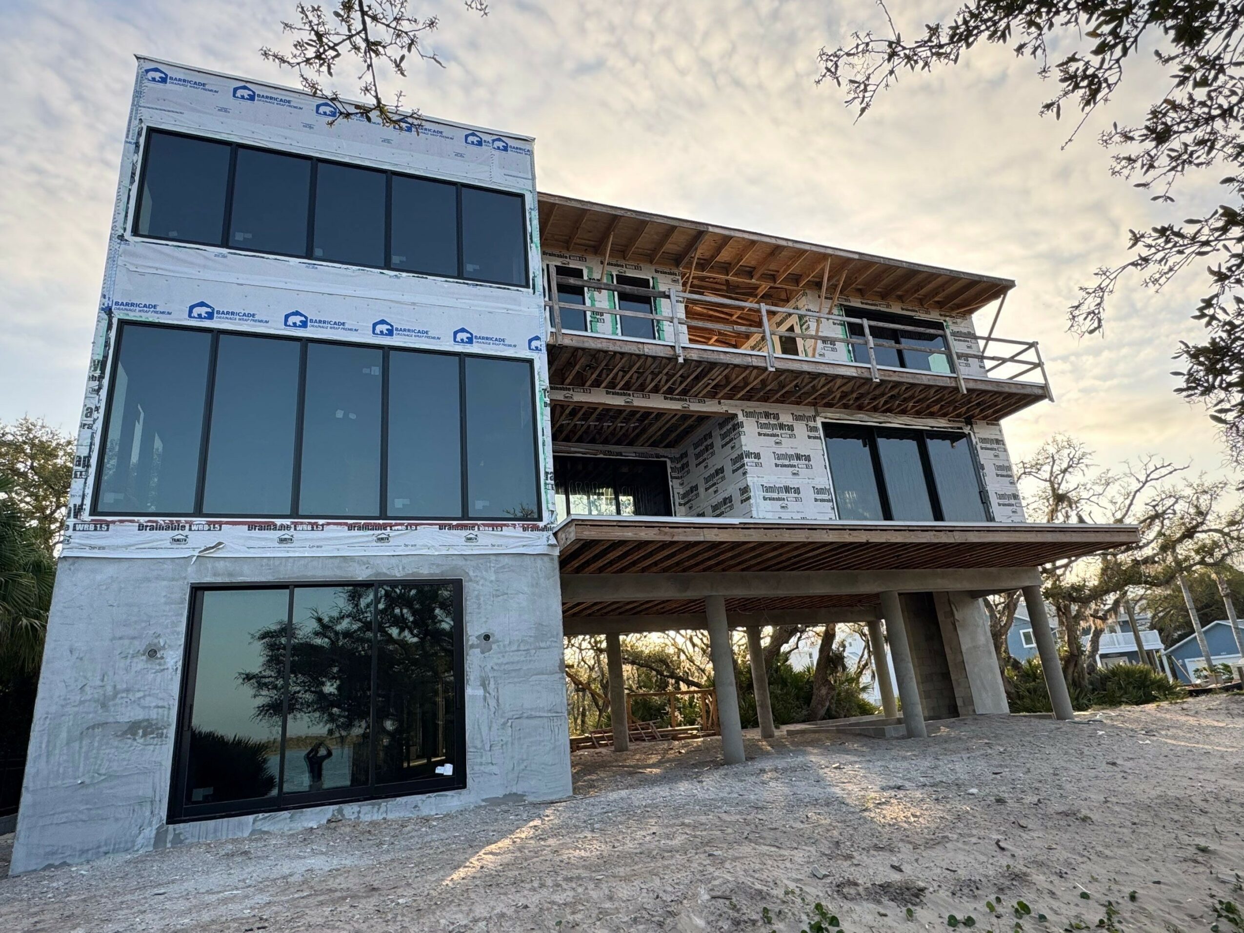 Building under construction with large windows, multiple floors, and an open ground level, set outdoors with trees and sky visible.