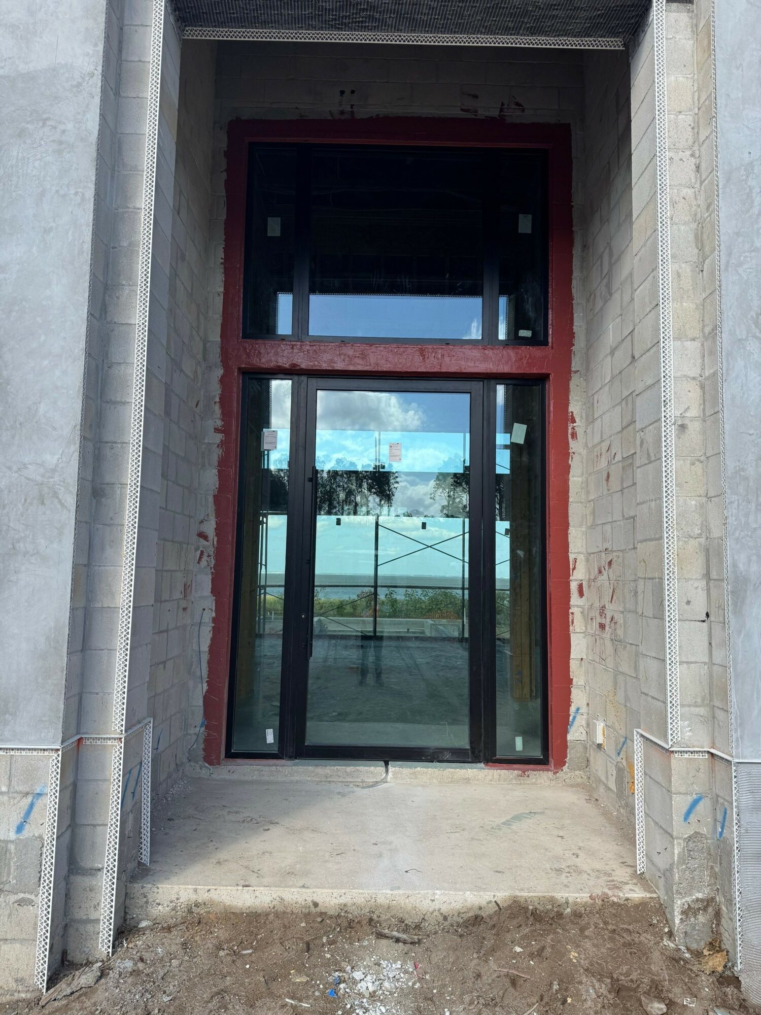 A glass door with a red frame set in a concrete wall, reflecting a landscape with trees and sky.