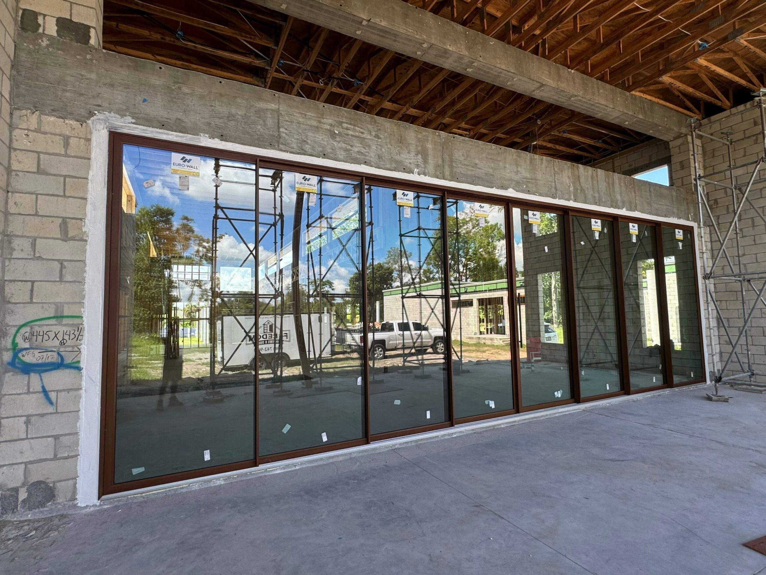 Large glass window with multiple panels reflecting trees and sky, set in a concrete wall under construction.