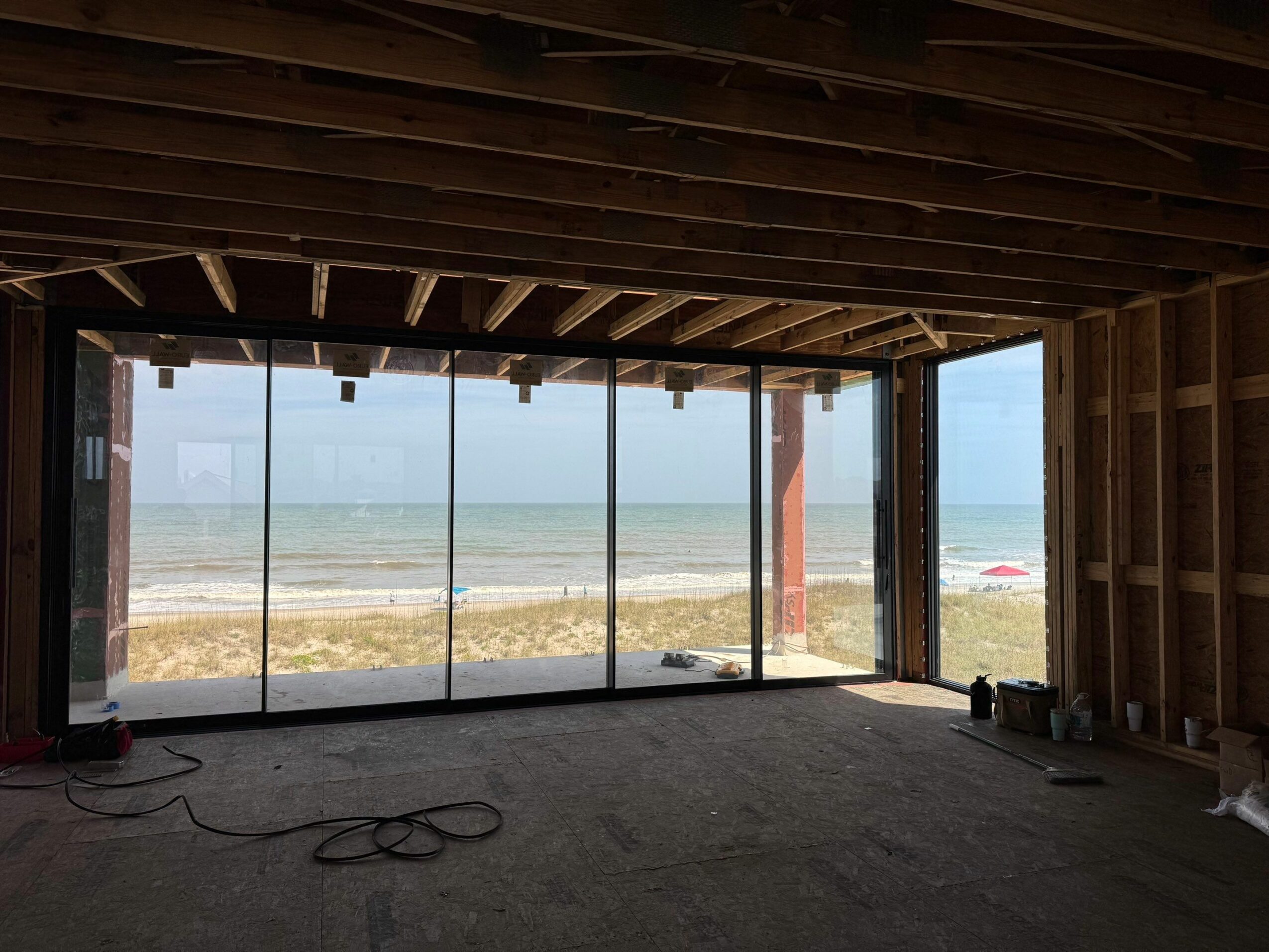 Interior view of a room under construction with large glass windows overlooking a beach and ocean.