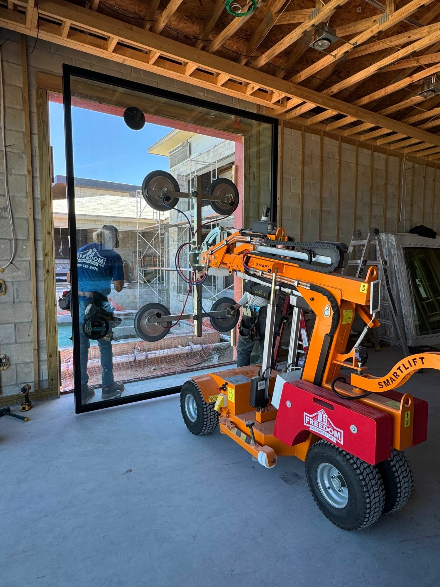 Construction site with a large glass window and a robotic lift in front, under an unfinished ceiling with exposed wooden beams.