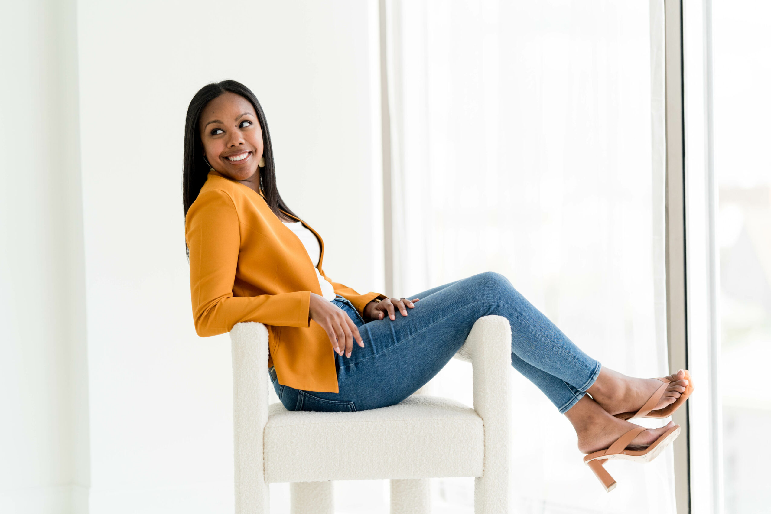 Woman sitting on a white chair near a large window, smiling, wearing a yellow top and blue jeans.