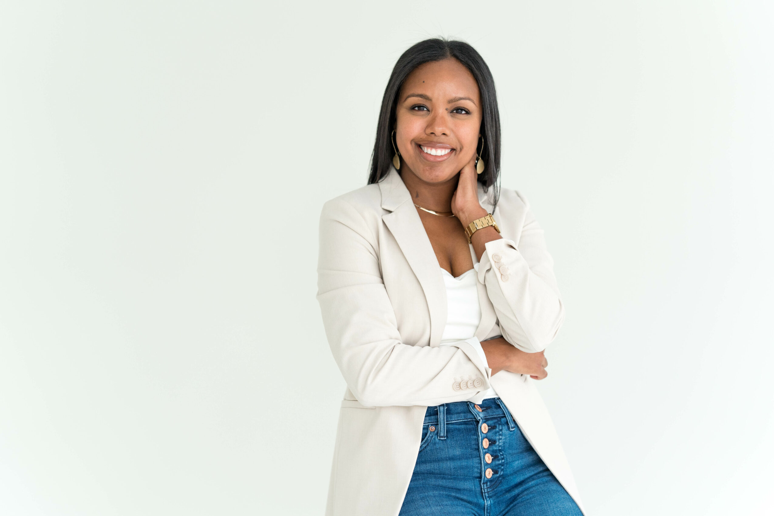 Smiling woman with long dark hair wearing a white blazer and blue jeans, standing against a light background.