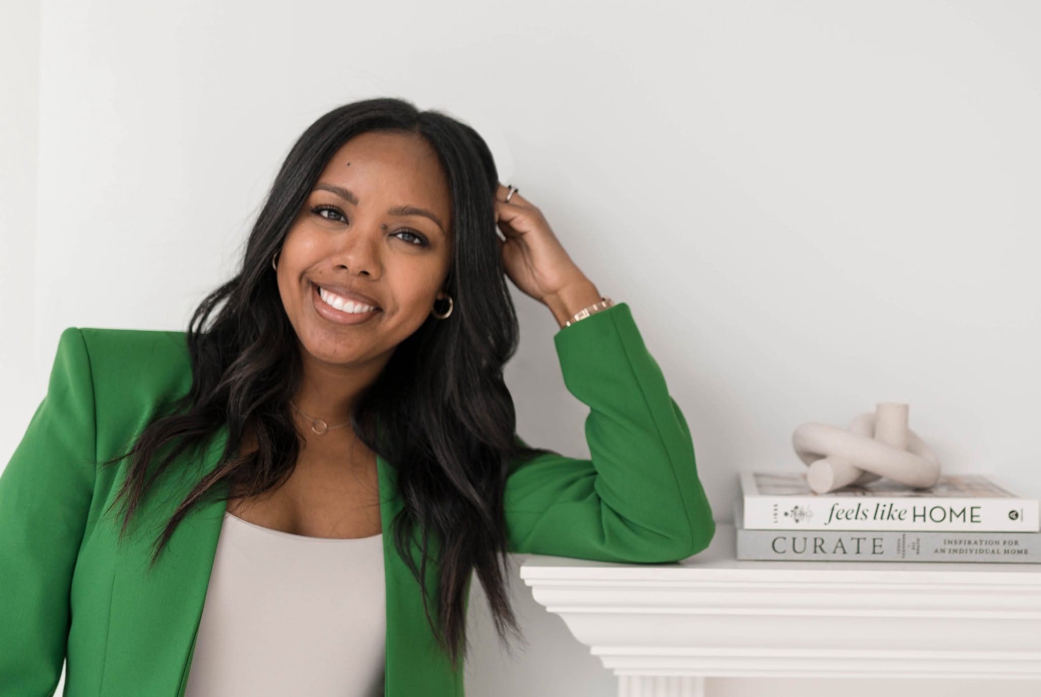 Smiling woman with long dark hair wearing a green blazer, sitting beside a white shelf with books and decor.