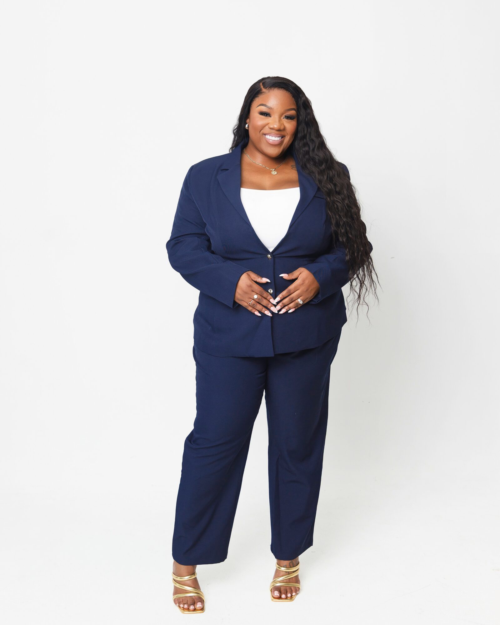 Smiling woman in a navy suit with white top, standing against a white background, hands clasped, long curly hair, gold sandals.