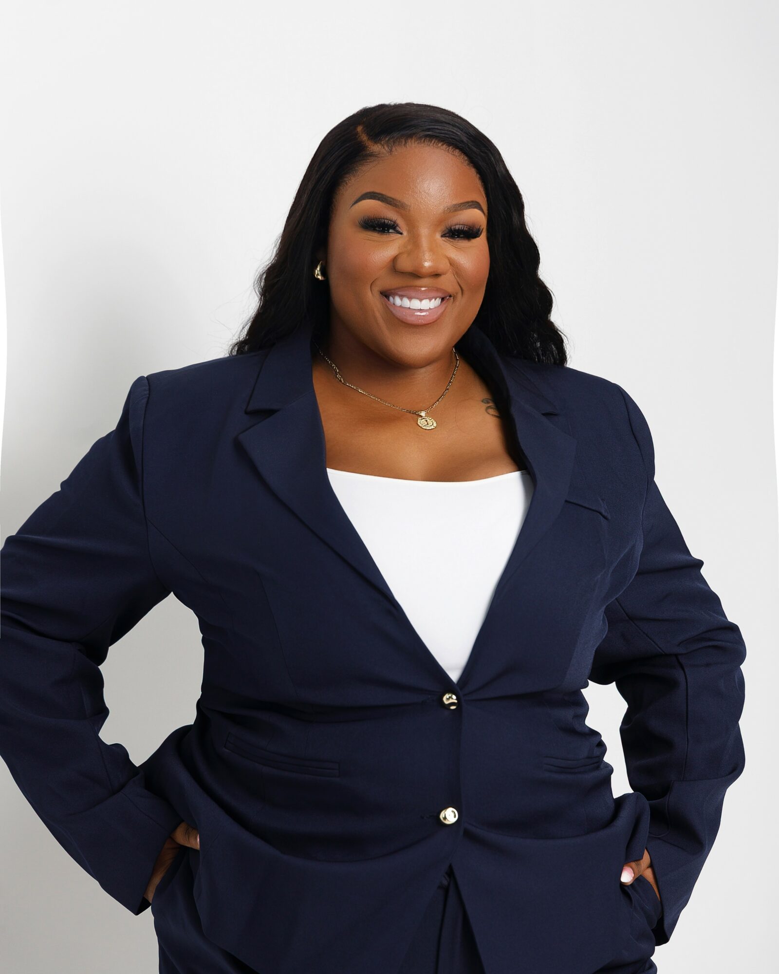 Smiling woman in navy blazer and white top poses with hands in pockets against a plain background.