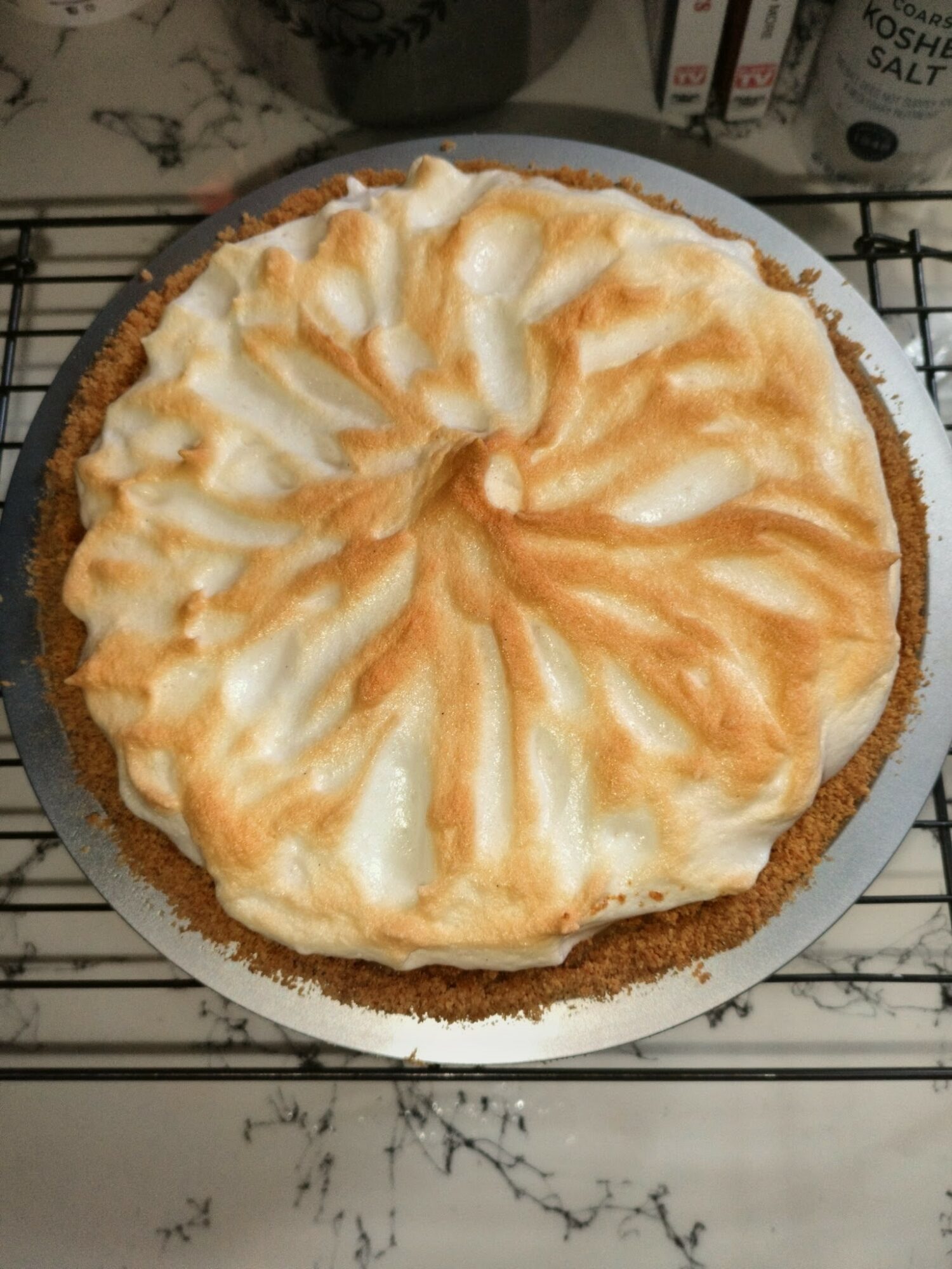 A baked pie with a golden crust and a creamy, swirled topping on a white plate, placed on a black wire rack.