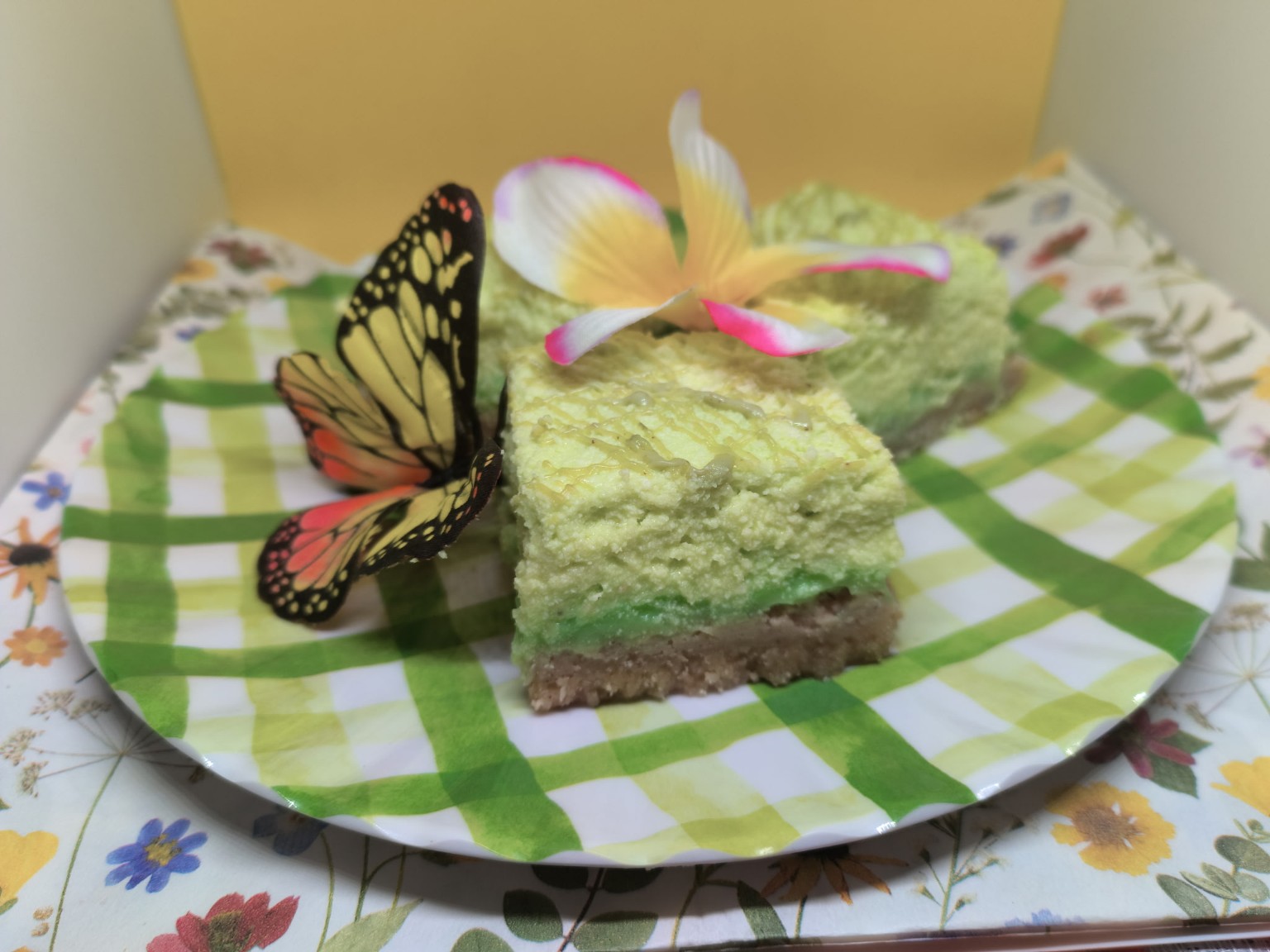 Two green layered desserts with pink and white flowers and a butterfly on a checkered paper plate, on a floral tablecloth.