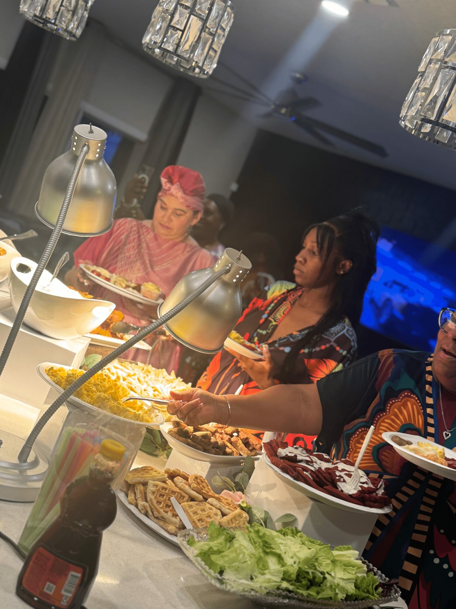 Two women and a child at a buffet table with various dishes, lettuce, and condiments, in a well-lit room.