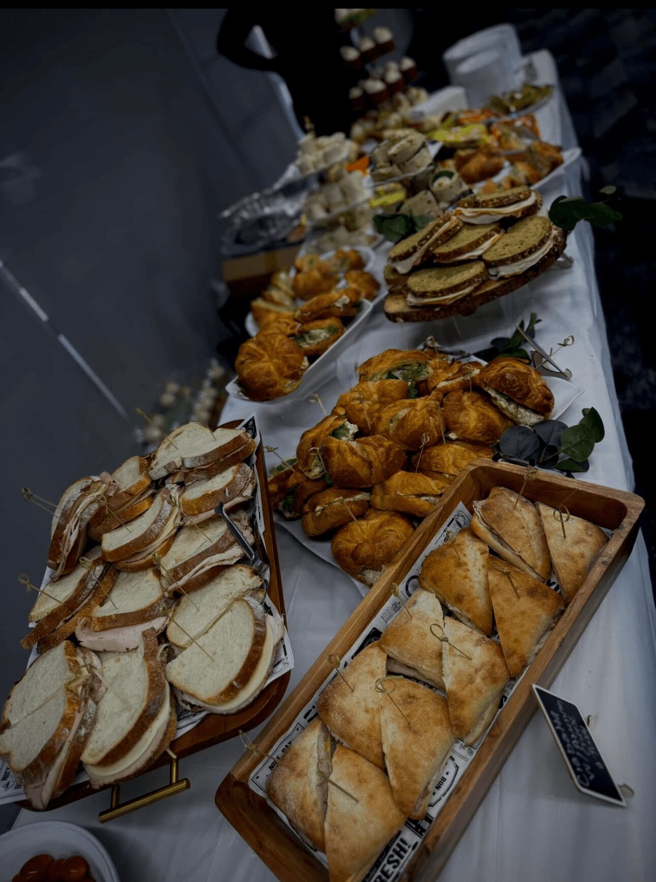 Table with various sandwiches, pastries, and finger foods arranged on plates and trays, with a blurred background.