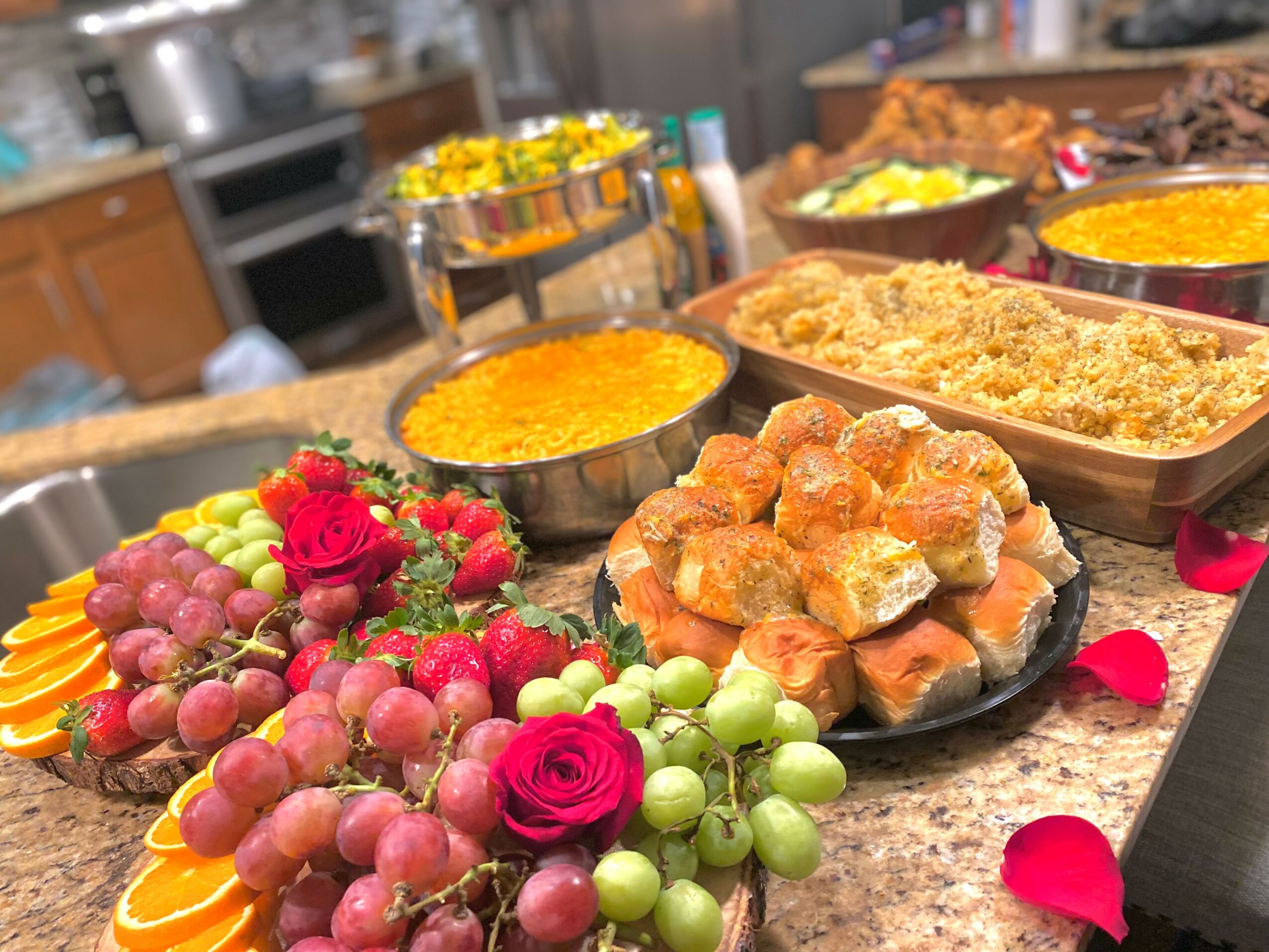 Assorted desserts and fruits on a kitchen counter, including grapes, roses, and baked goods, with a blurred background.