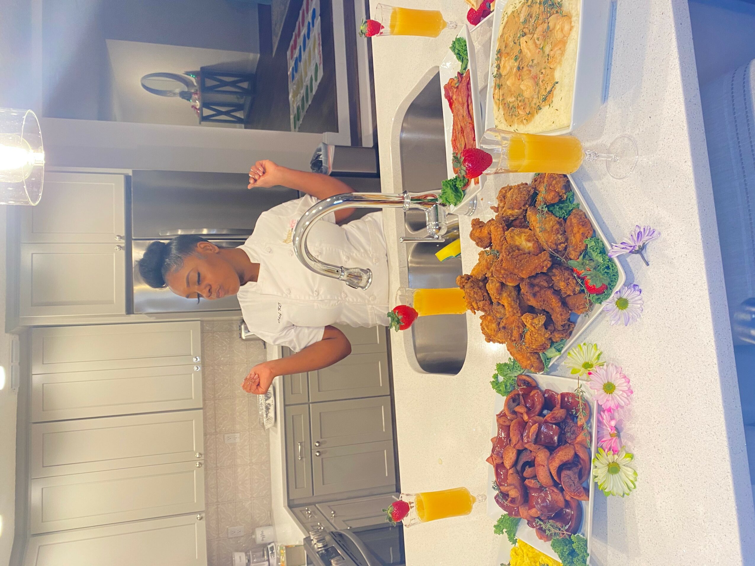 Person in a white shirt standing in a kitchen with a table of fried chicken, shrimp, and drinks, decorated with candles and flowers.