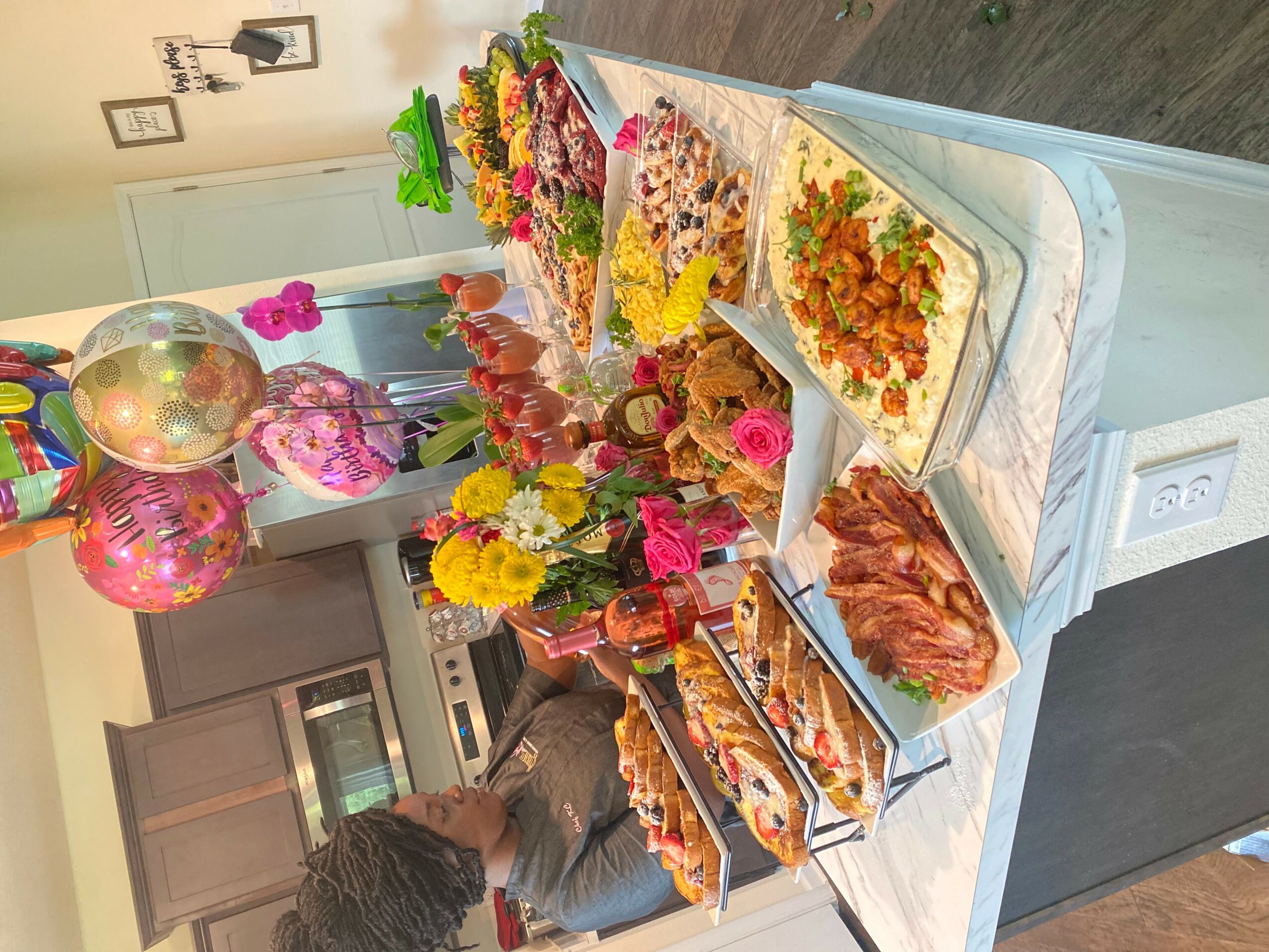 Table with various dishes, flowers, and decorations in a kitchen setting, with a person reaching for food.