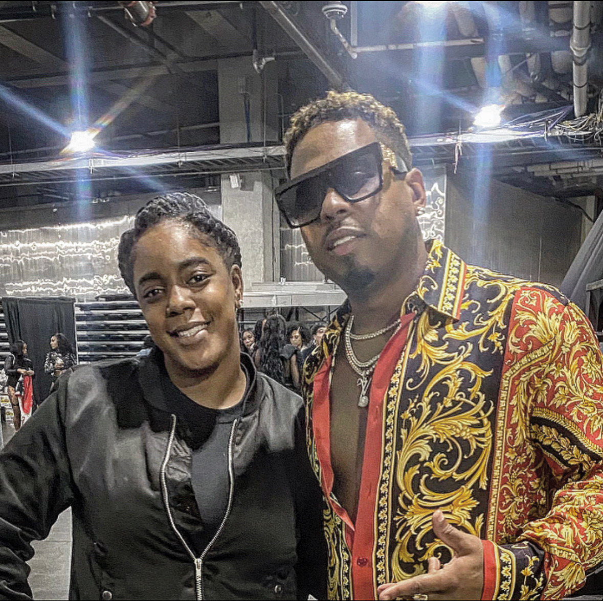 Two people posing indoors, one wearing sunglasses and a patterned shirt, the other in a black jacket, smiling.