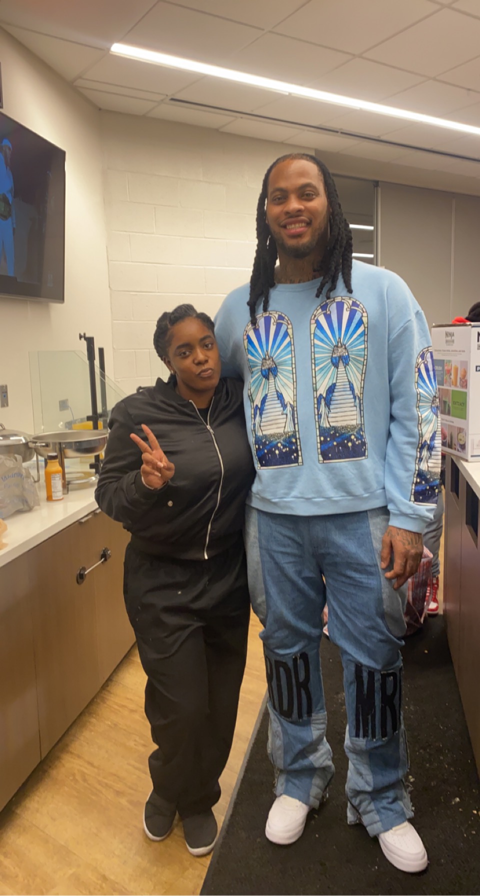 Two people standing in a room, one taller with long hair and a blue outfit, the other shorter with dark hair and a black outfit.