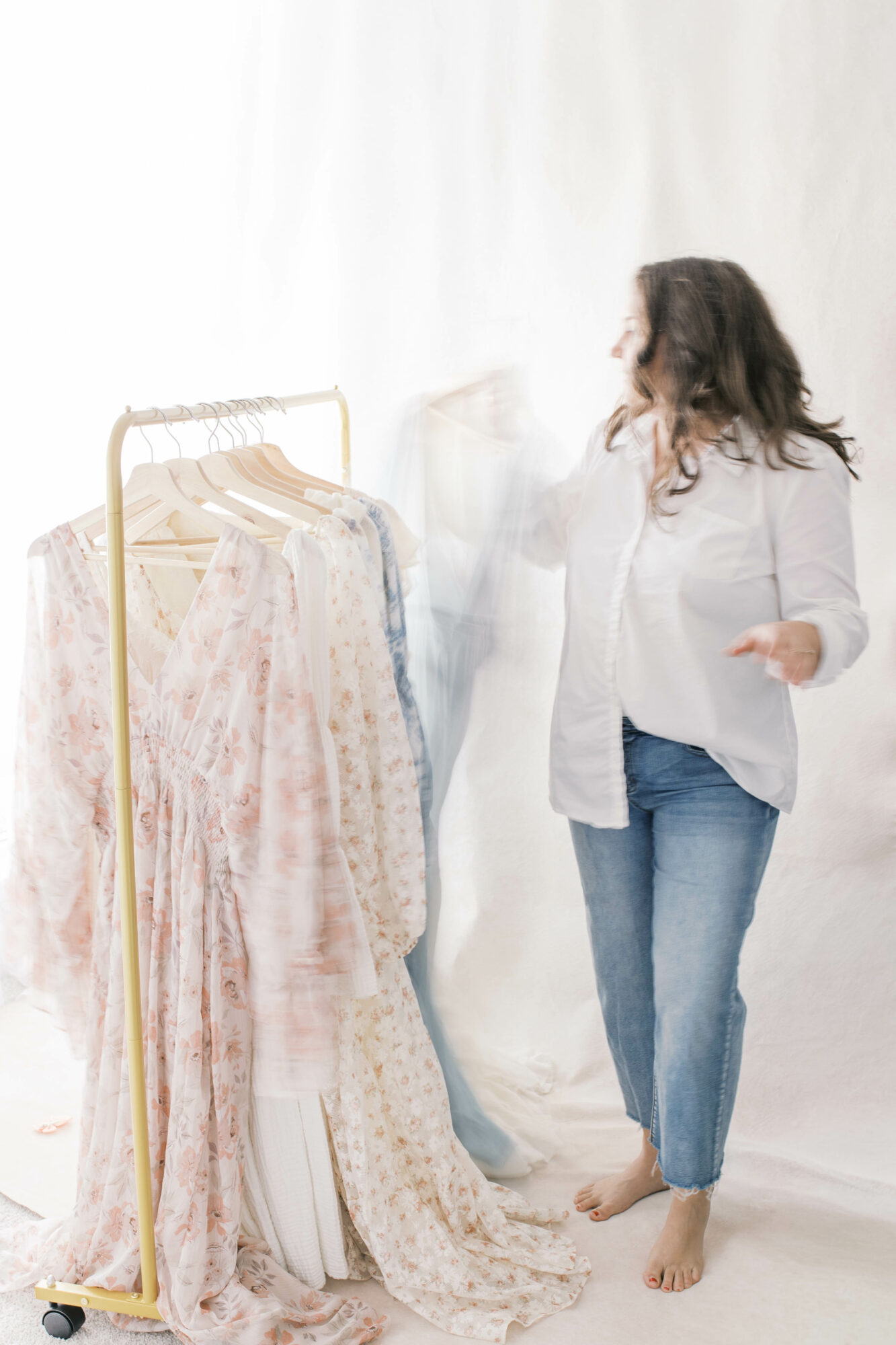 Woman with long hair wearing a white shirt and jeans standing near a clothing rack with dresses, on a white background.