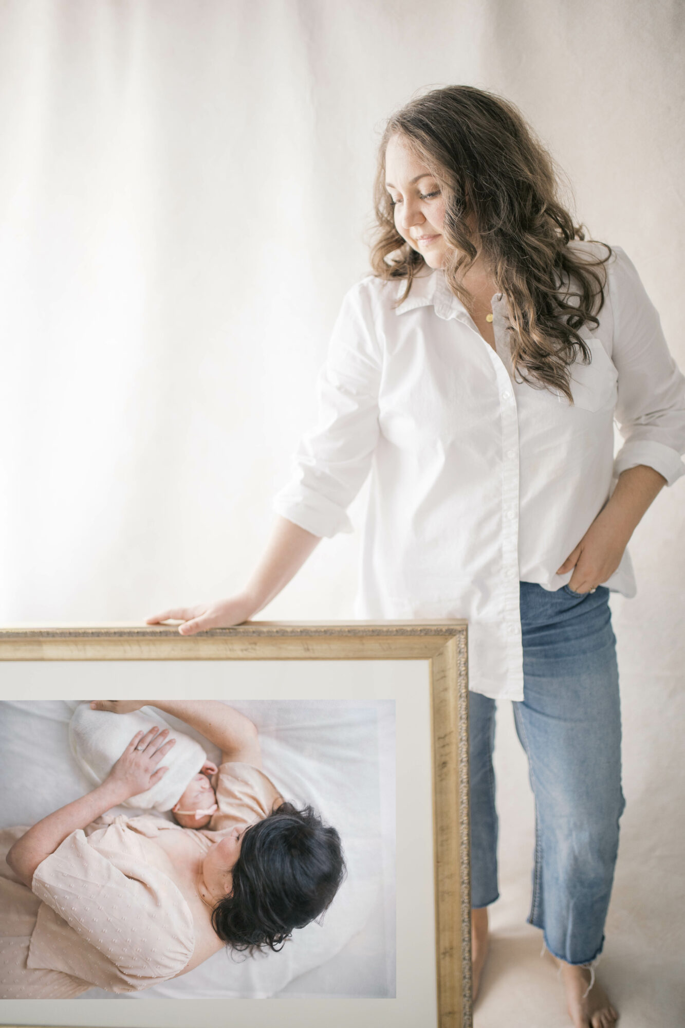 Woman with long curly hair in a white shirt and jeans stands next to a framed photo of a woman lying down, on a light background.