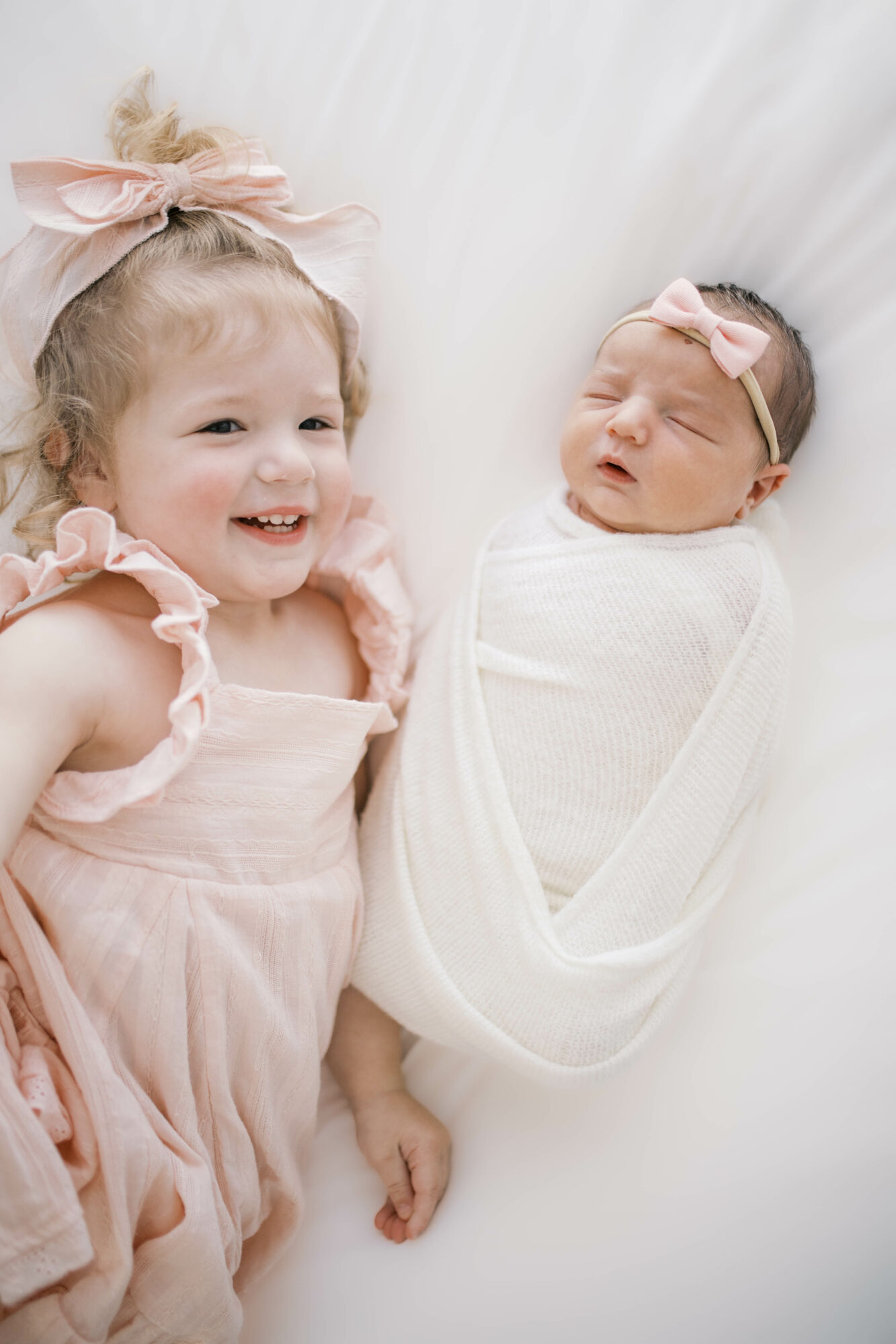 A young girl with curly hair and a pink dress smiling next to a newborn wrapped in white swaddle, both with pink bows.